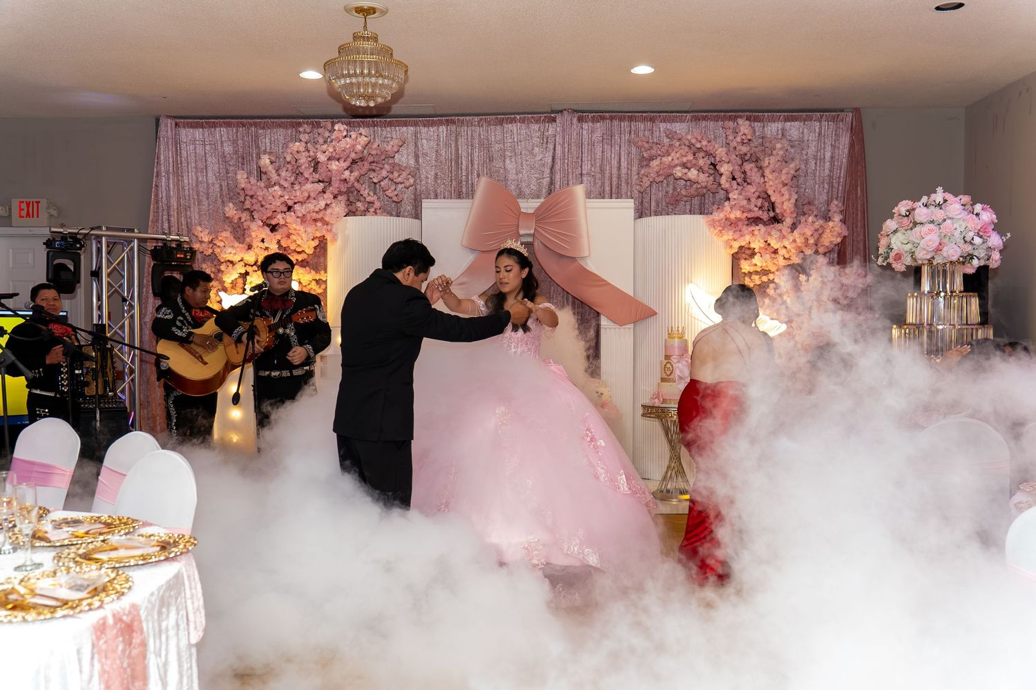 Quinceañera celebration with live mariachi band, girl in pink ball gown dancing in fog effect at decorated venue.