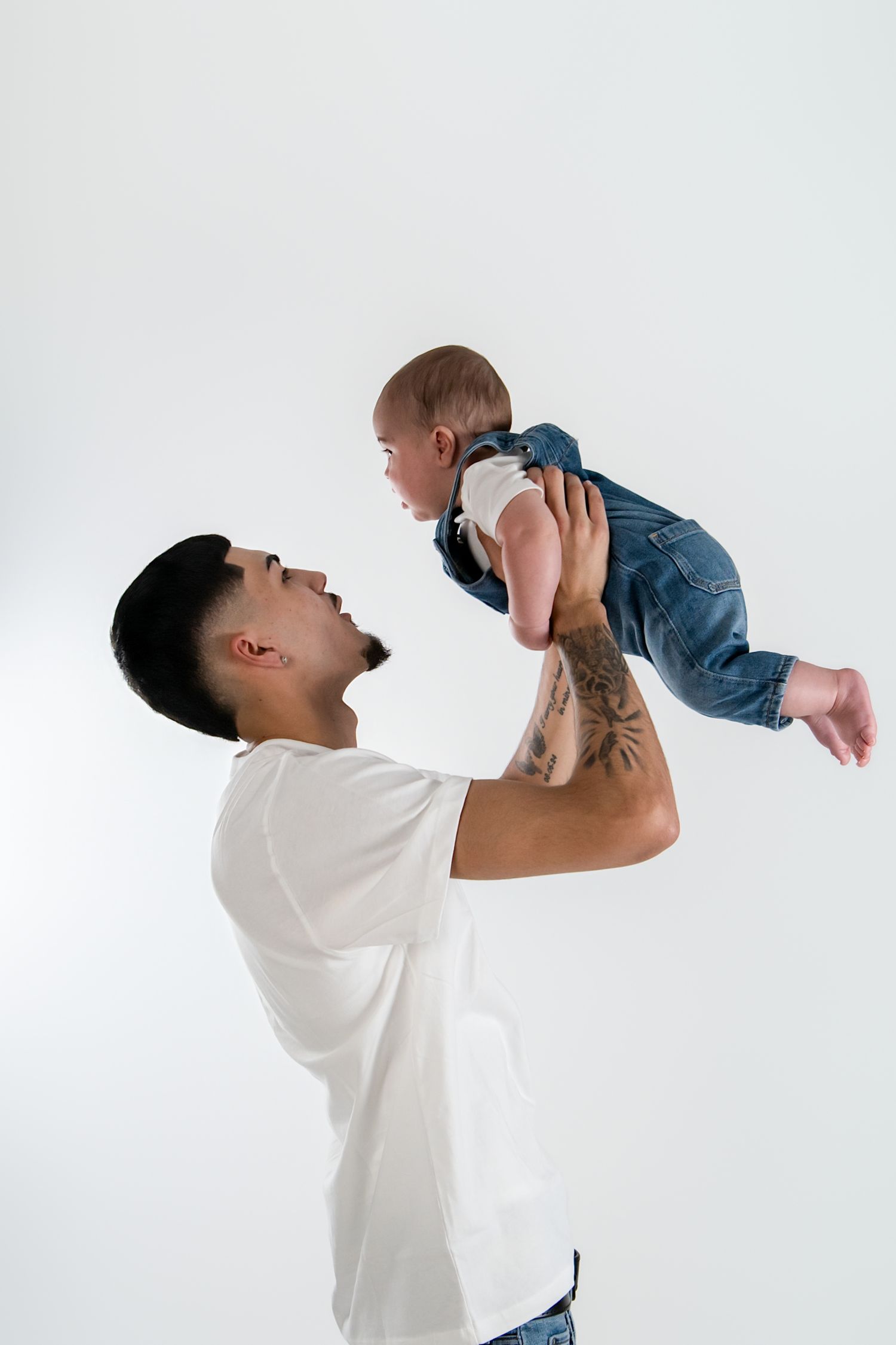 A tattooed father joyfully lifts his baby in the air against a white background. baby photoshoot