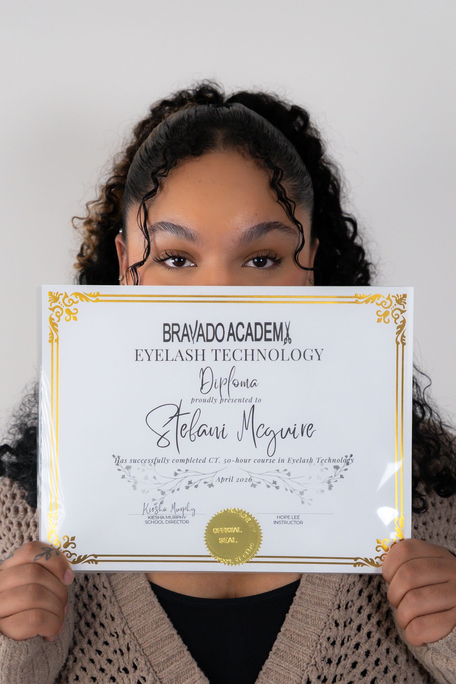 Young woman proudly holds Bravado Academy Eyelash Technology diploma certificate in studio setting.