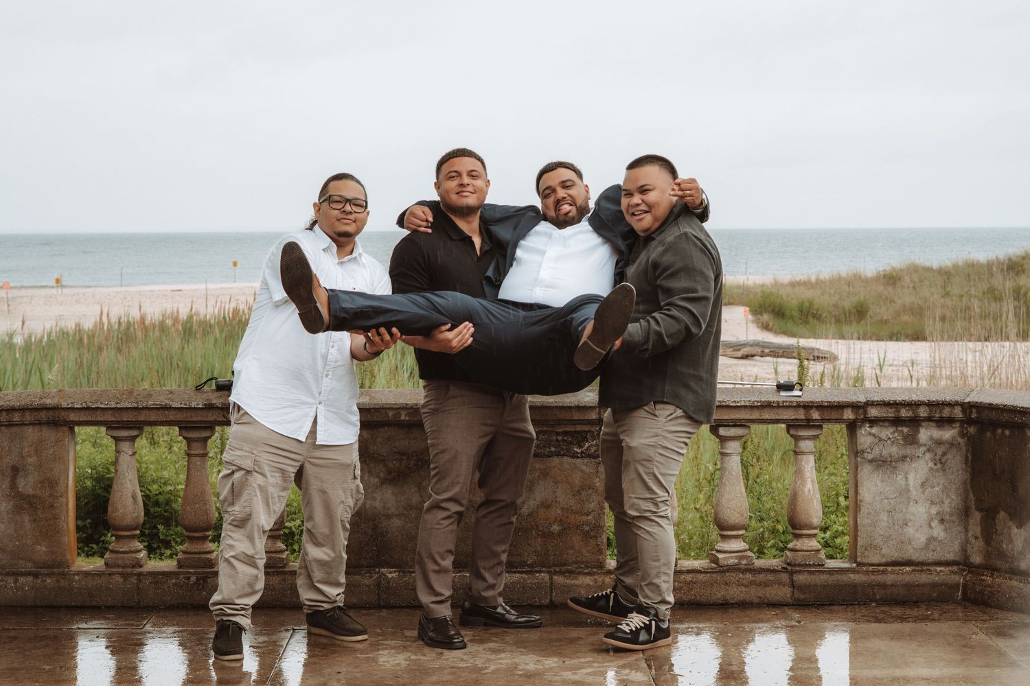 Four groomsmen celebrating at beach wedding venue, playfully lifting one man in the air on a cloudy day.