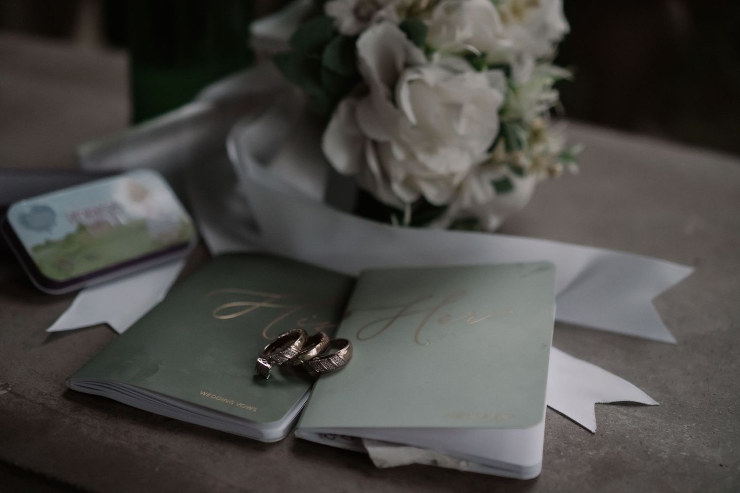 Wedding rings resting on sage green vow books beside a white floral bouquet and decorative tin.