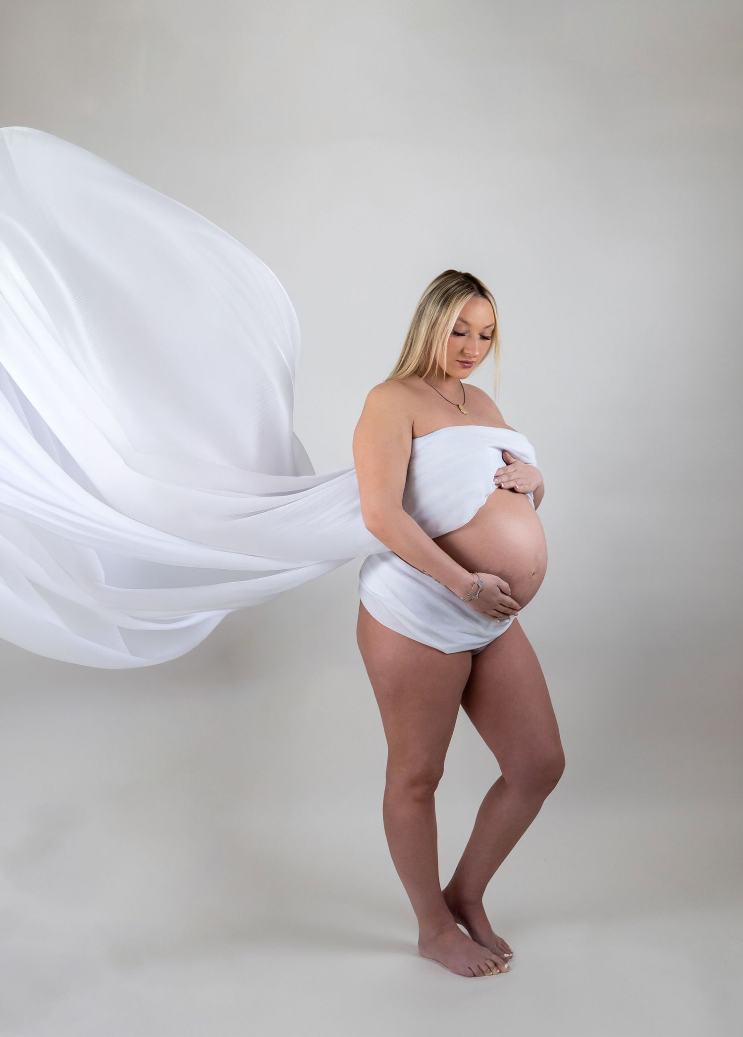 Pregnant woman wrapped in flowing white fabric cradling baby bump in studio portrait.