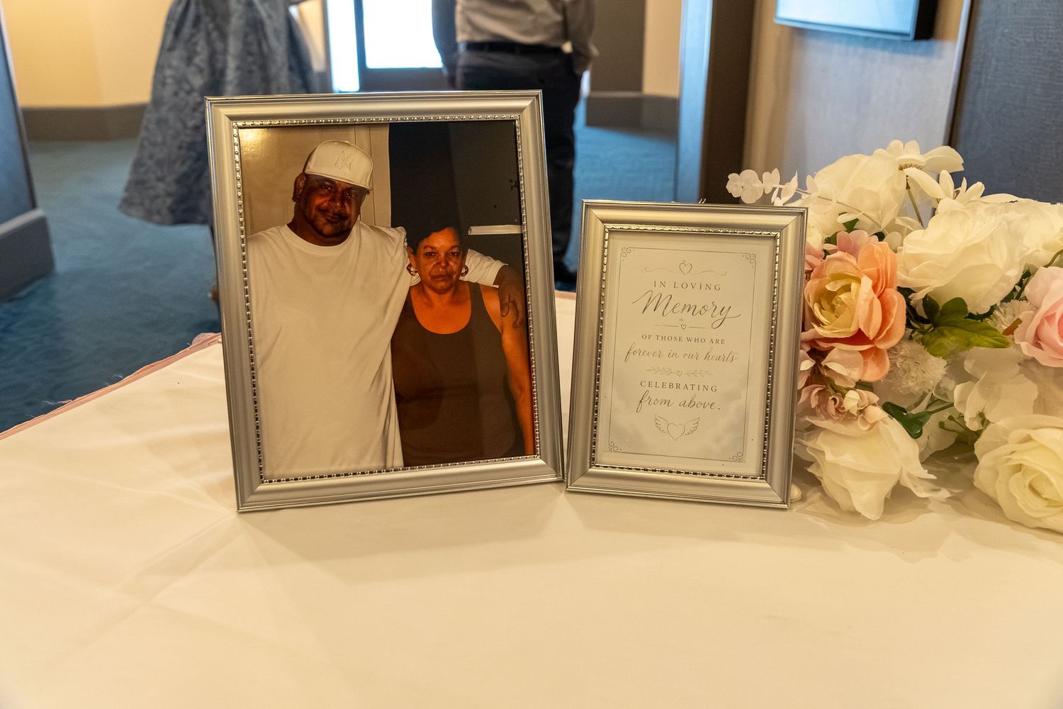 Memorial table display with framed photo of couple and In Memory sign beside pink and white floral arrangement. Sweet 16/Quinceañera