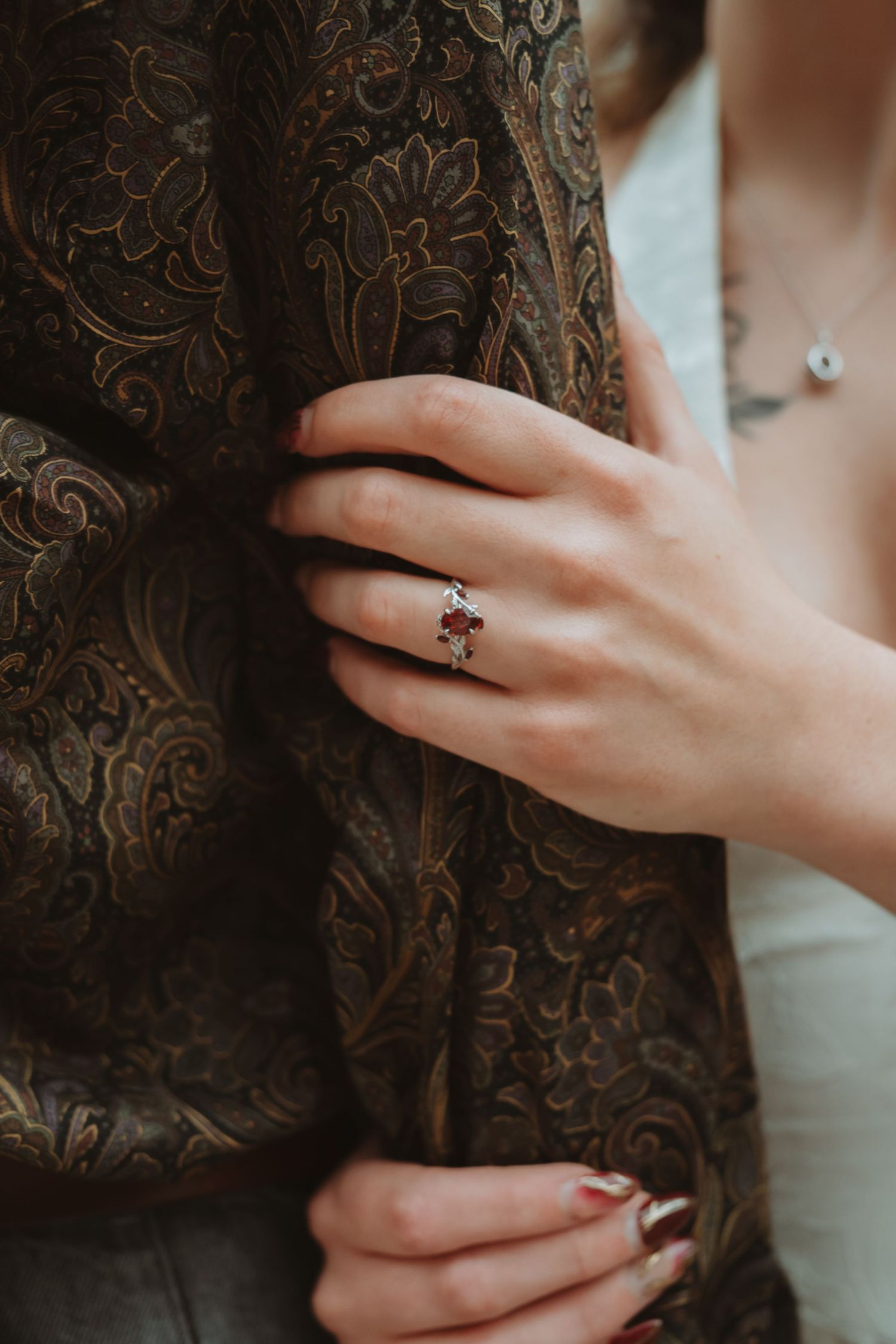 Woman's hand with red nail polish wearing a vintage-style red gemstone ring, gripping a dark paisley fabric jacket.