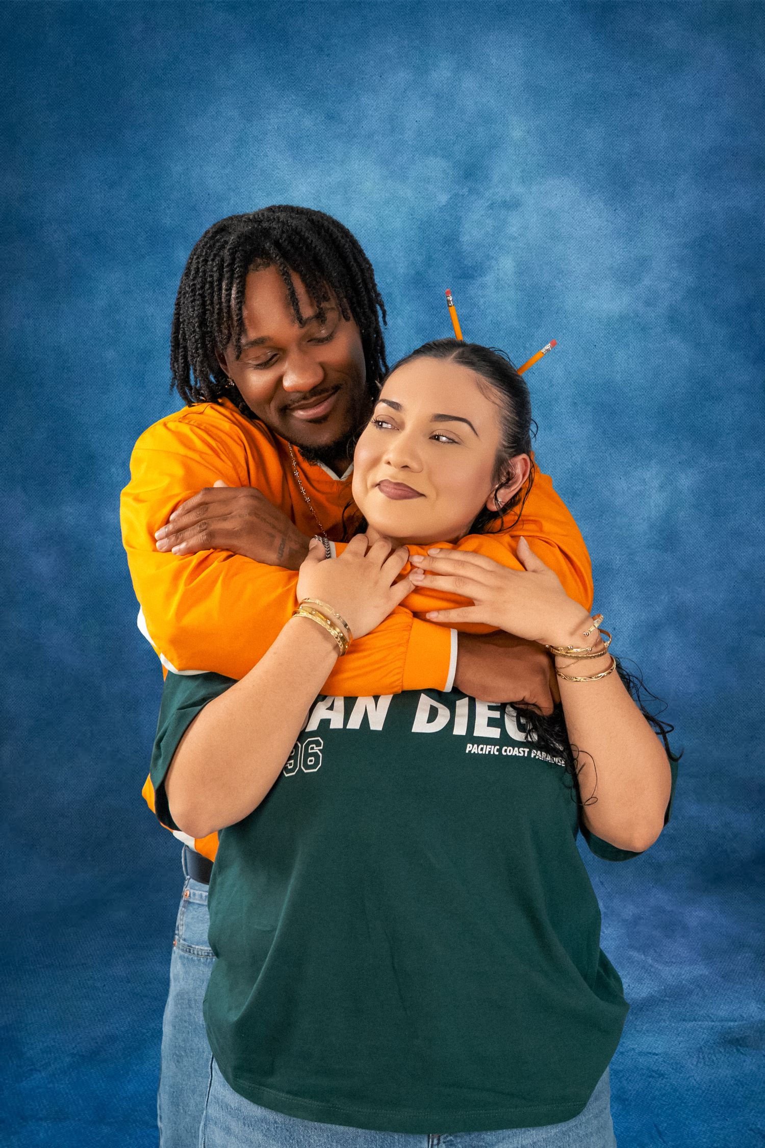 Smiling couple embracing against a blue studio backdrop, one in an orange jacket hugging the other in a green San Diego sweatshirt.