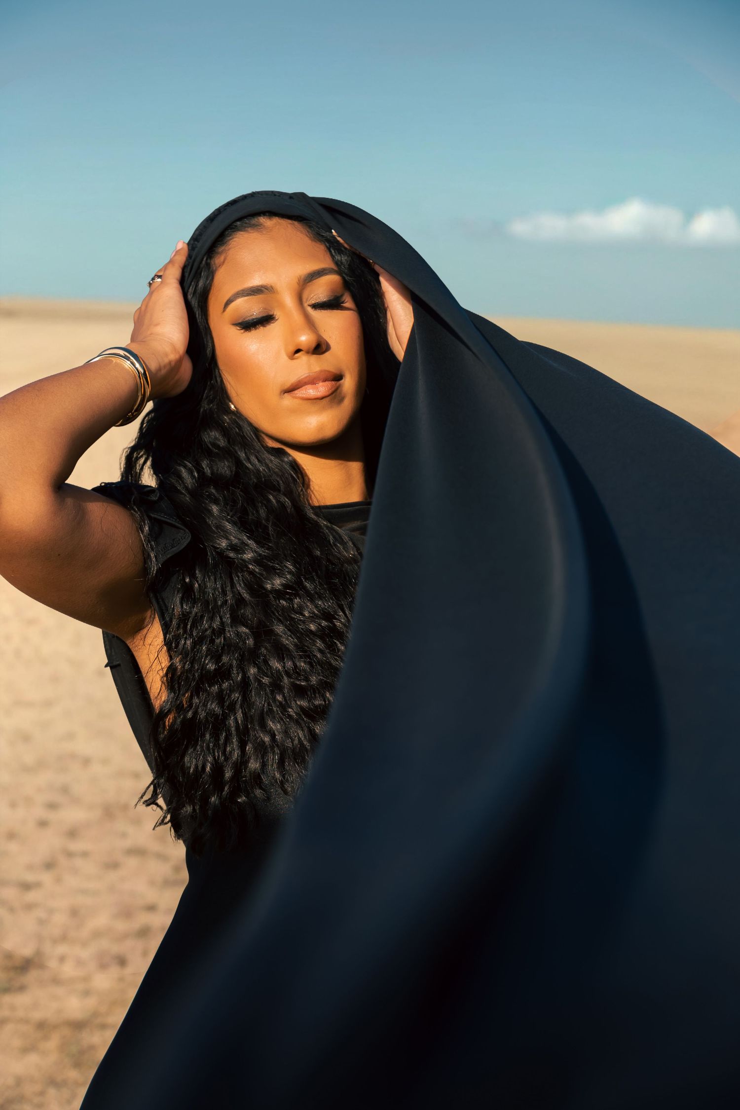 A woman with long black hair poses in a desert, draped in flowing black fabric under a clear blue sky.