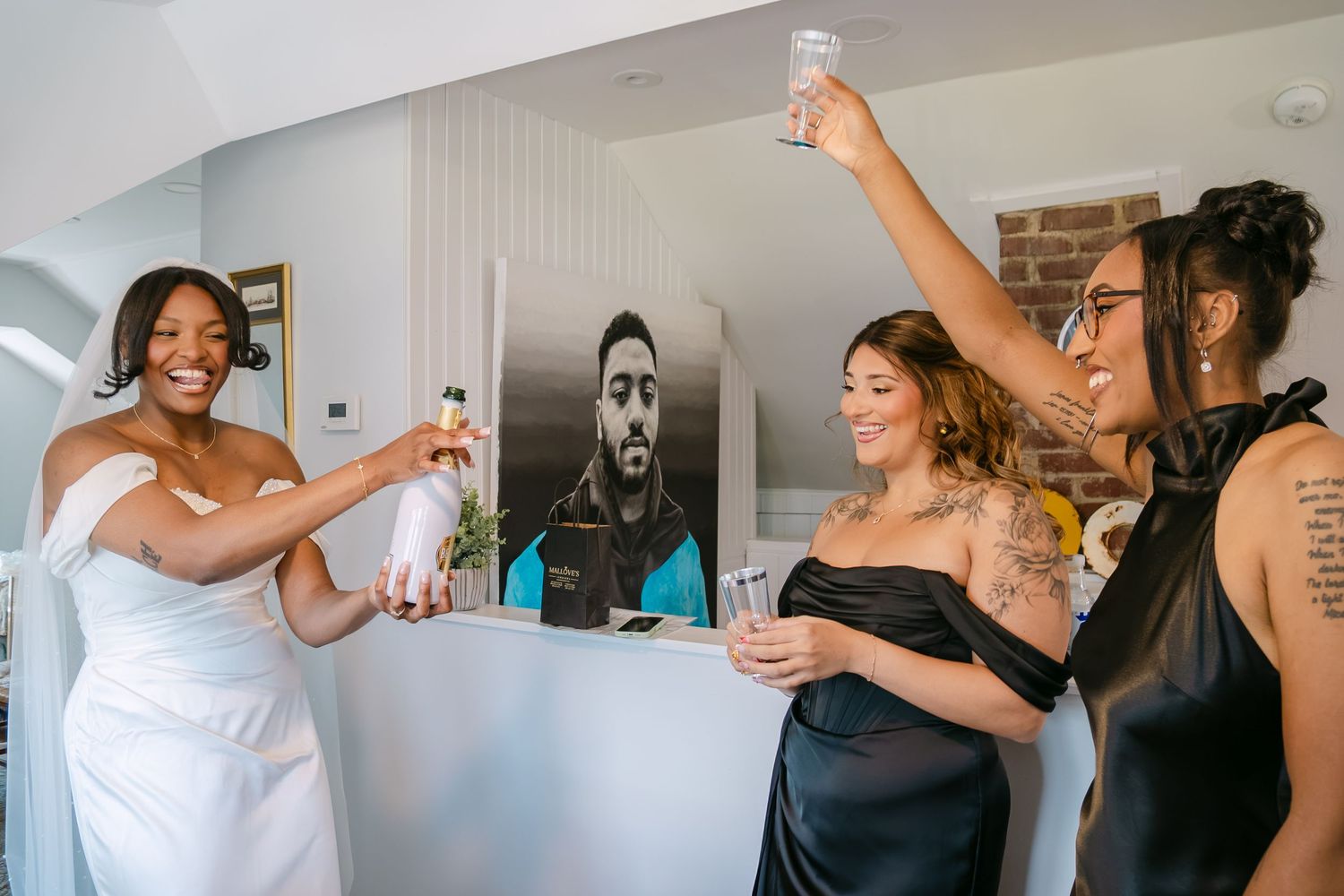Bride in white gown toasts with two bridesmaids in black dresses, celebrating together in a modern kitchen.