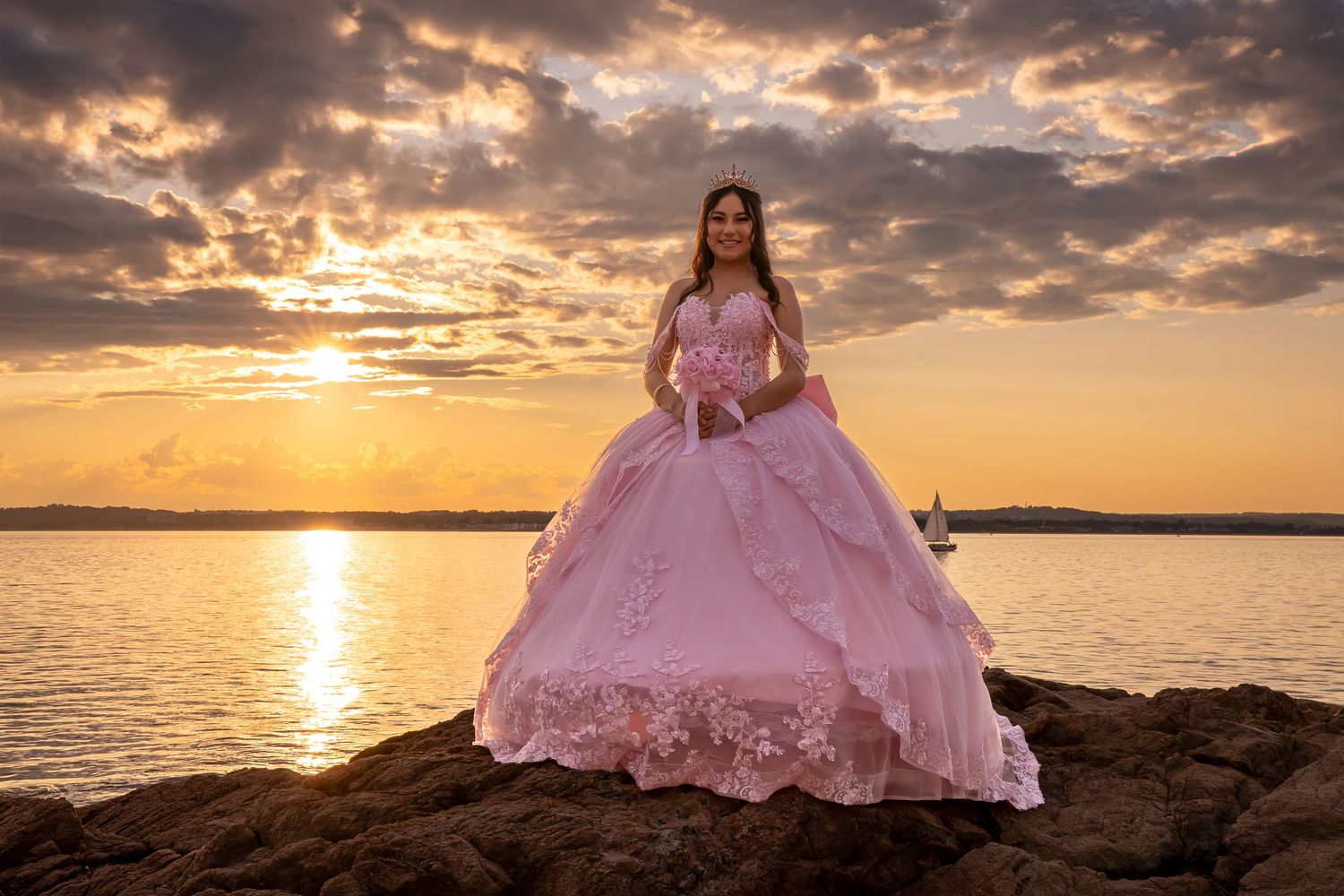 Young woman in pink ball gown poses on rocky shore at golden sunset over calm water.