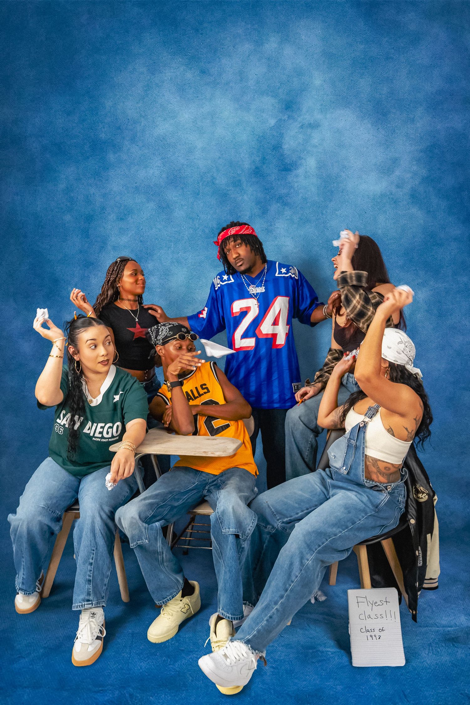 Five stylishly dressed friends in streetwear, jerseys and baggy jeans pose playfully around a school desk against a blue studio backdrop.