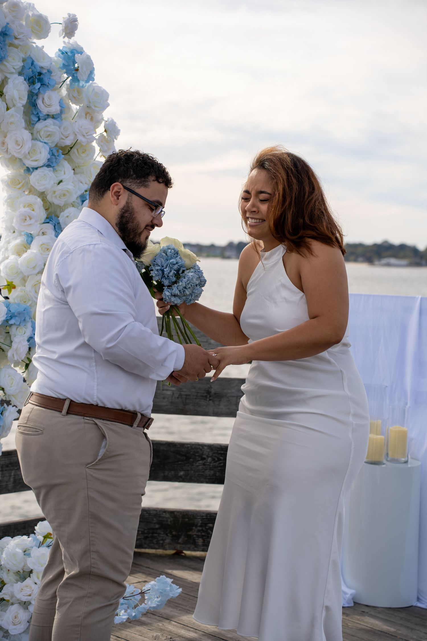 Smiling bride in a white satin dress holds a blue and white bouquet while exchanging rings with her groom at a waterfront wedding ceremony.