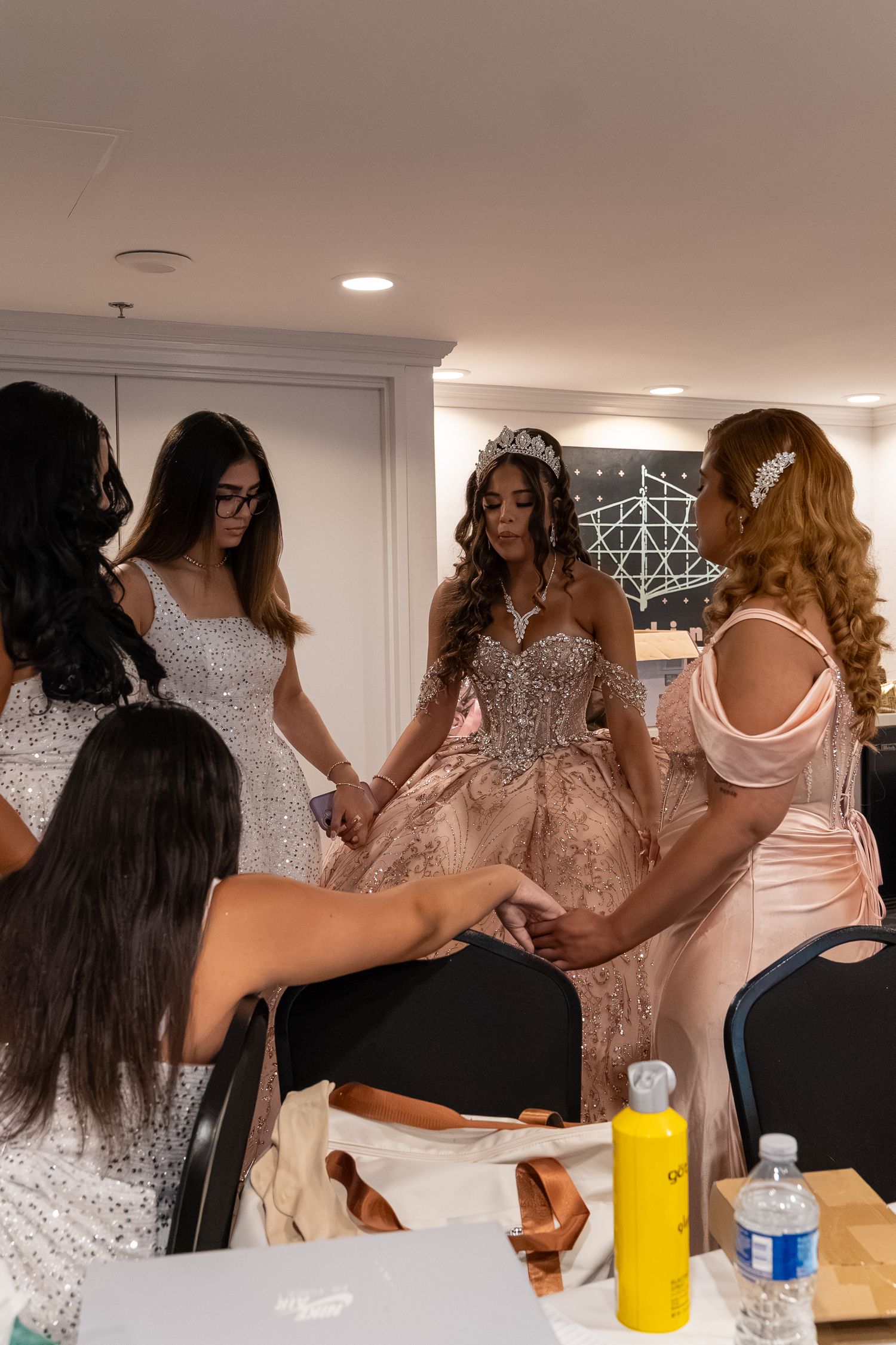Young woman in ornate pink quinceañera gown with tiara surrounded by attendants in preparation room. Sweet 16/Quinceañera