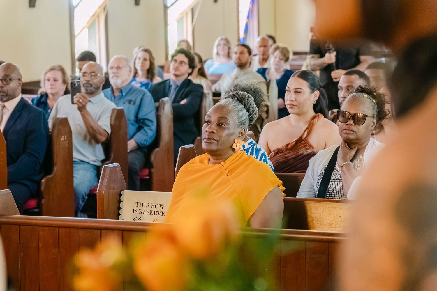 Congregation seated in wooden church pews during a ceremony, with a woman in yellow standing out among diverse attendees.