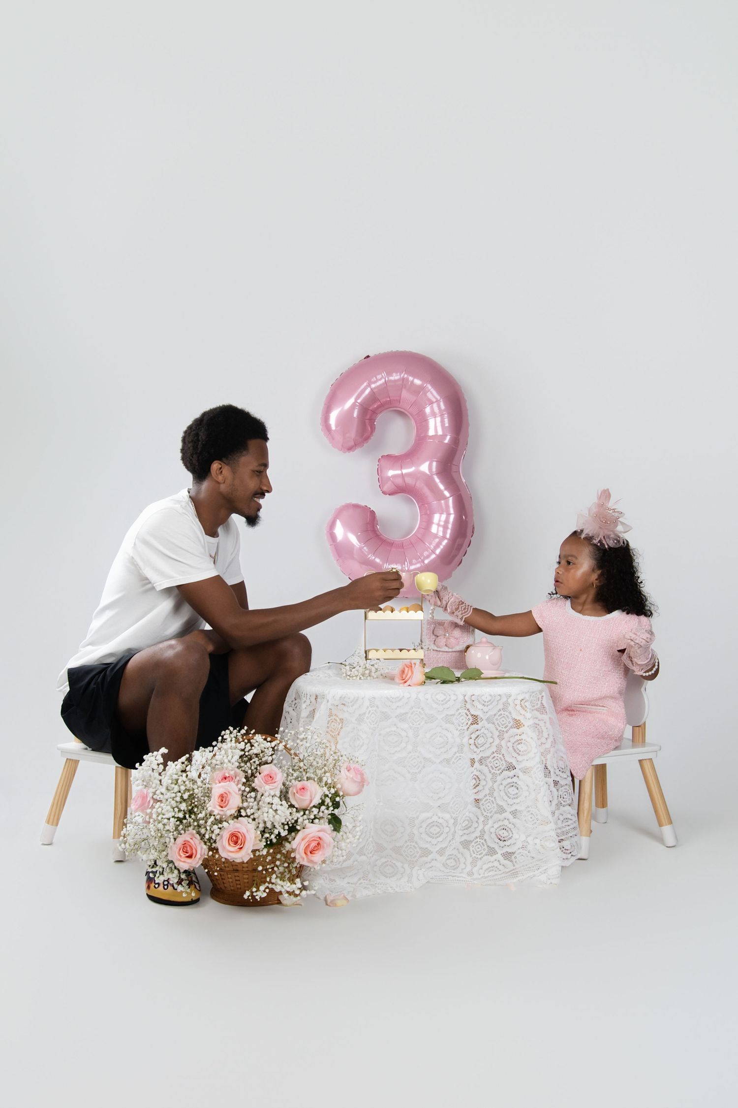 A father and daughter celebrate her 3rd birthday at a small table with cake, pink balloon, and flowers on white background.