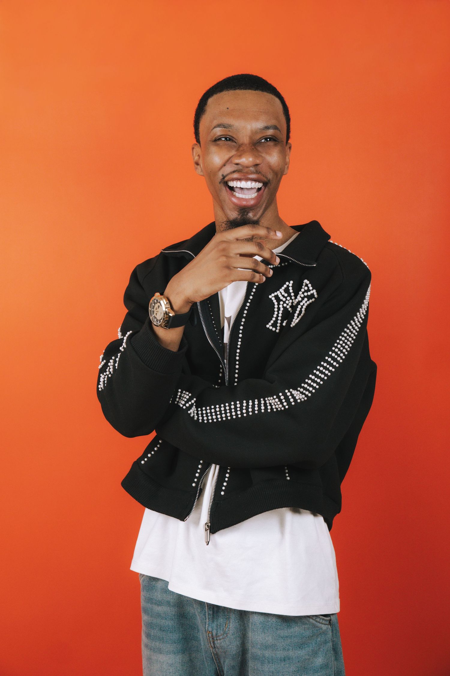 Smiling man in black studded jacket posing confidently against bright orange background.