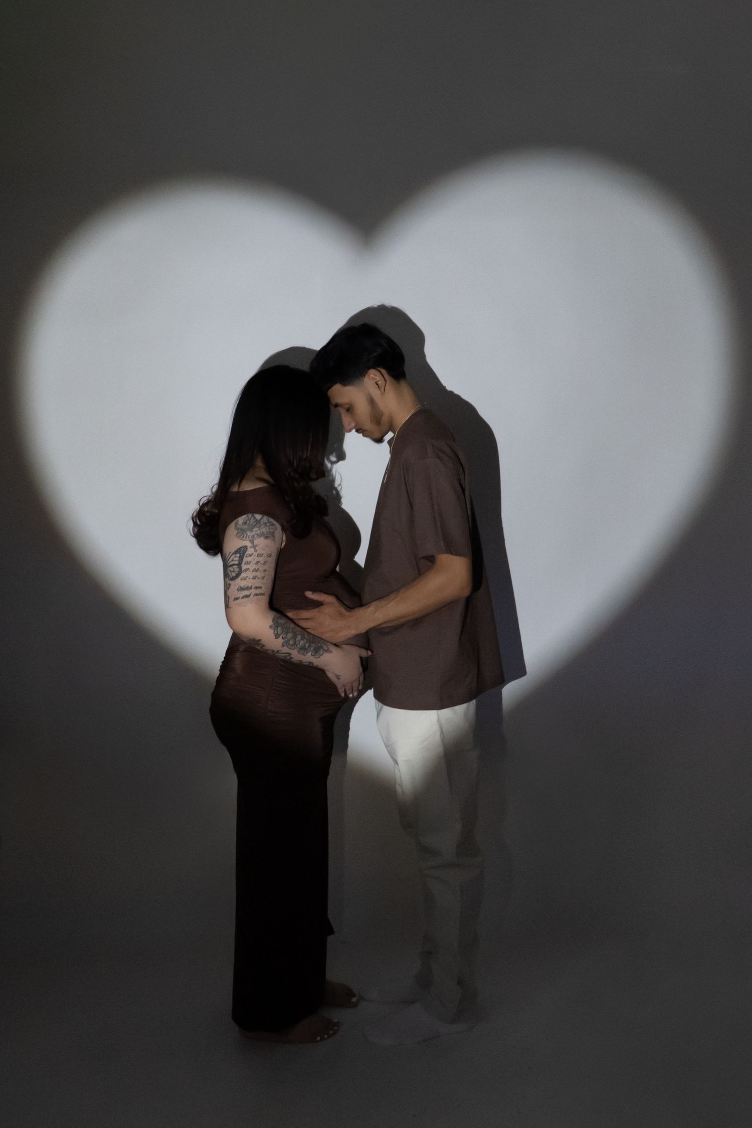 Couple tenderly touching pregnant belly framed by heart-shaped light projection on gray background.