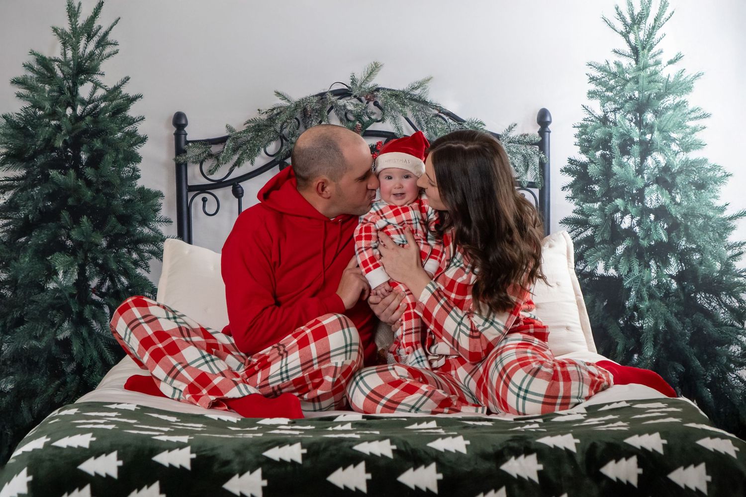 Family of three in matching red plaid pajamas sitting on bed with Christmas trees.