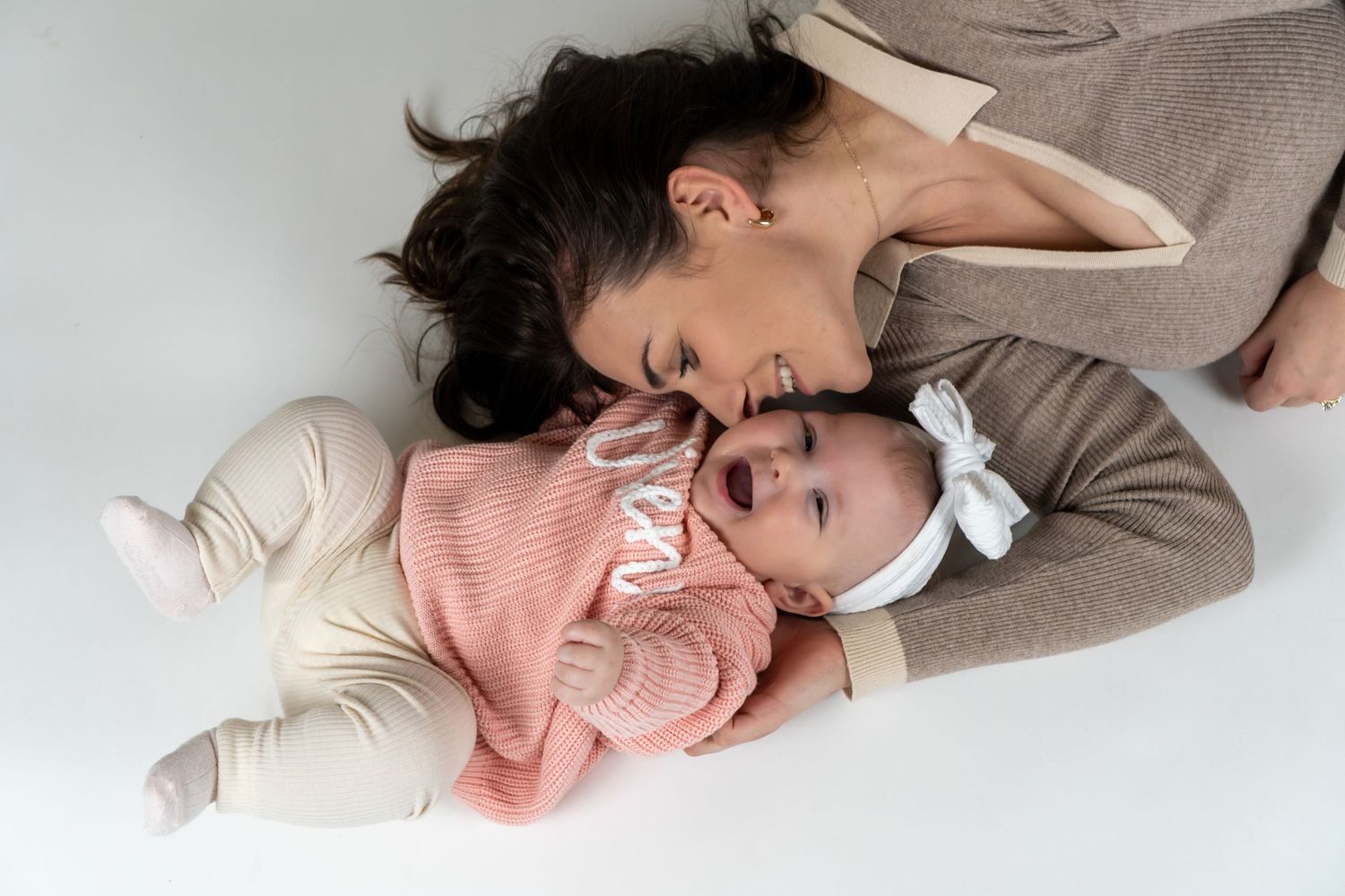 Loving mother lying on white floor kisses her newborn baby dressed in pink with a white bow headband.