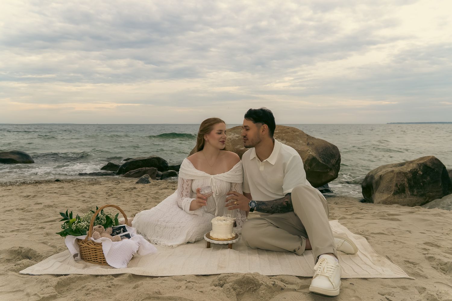 Couple sitting on a blanket at the beach during sunset, sharing a romantic picnic with wine and cake.