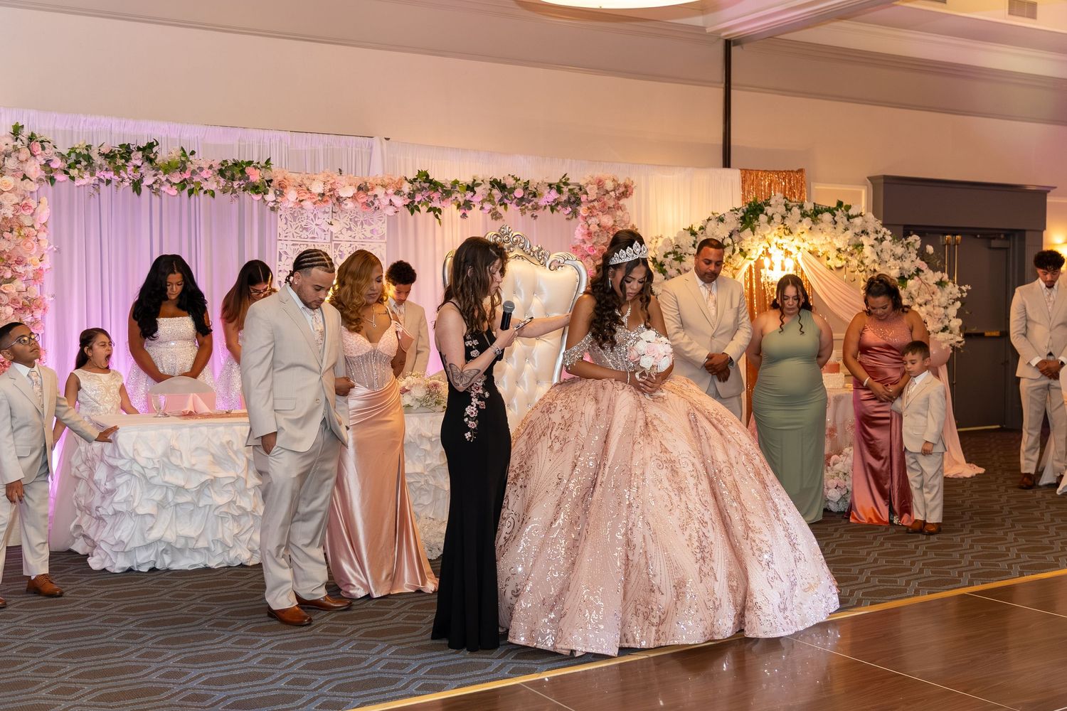 A young woman in a ornate pink ball gown celebrates her quinceañera surrounded by elegantly dressed guests. Sweet 16/Quinceañera