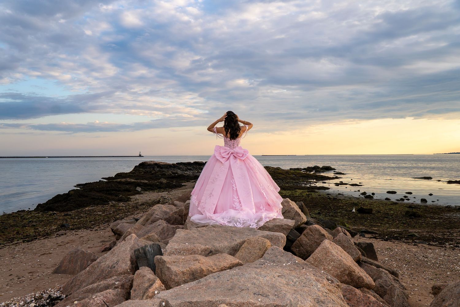 Girl in pink ball gown stands on rocky shore gazing at ocean sunset.