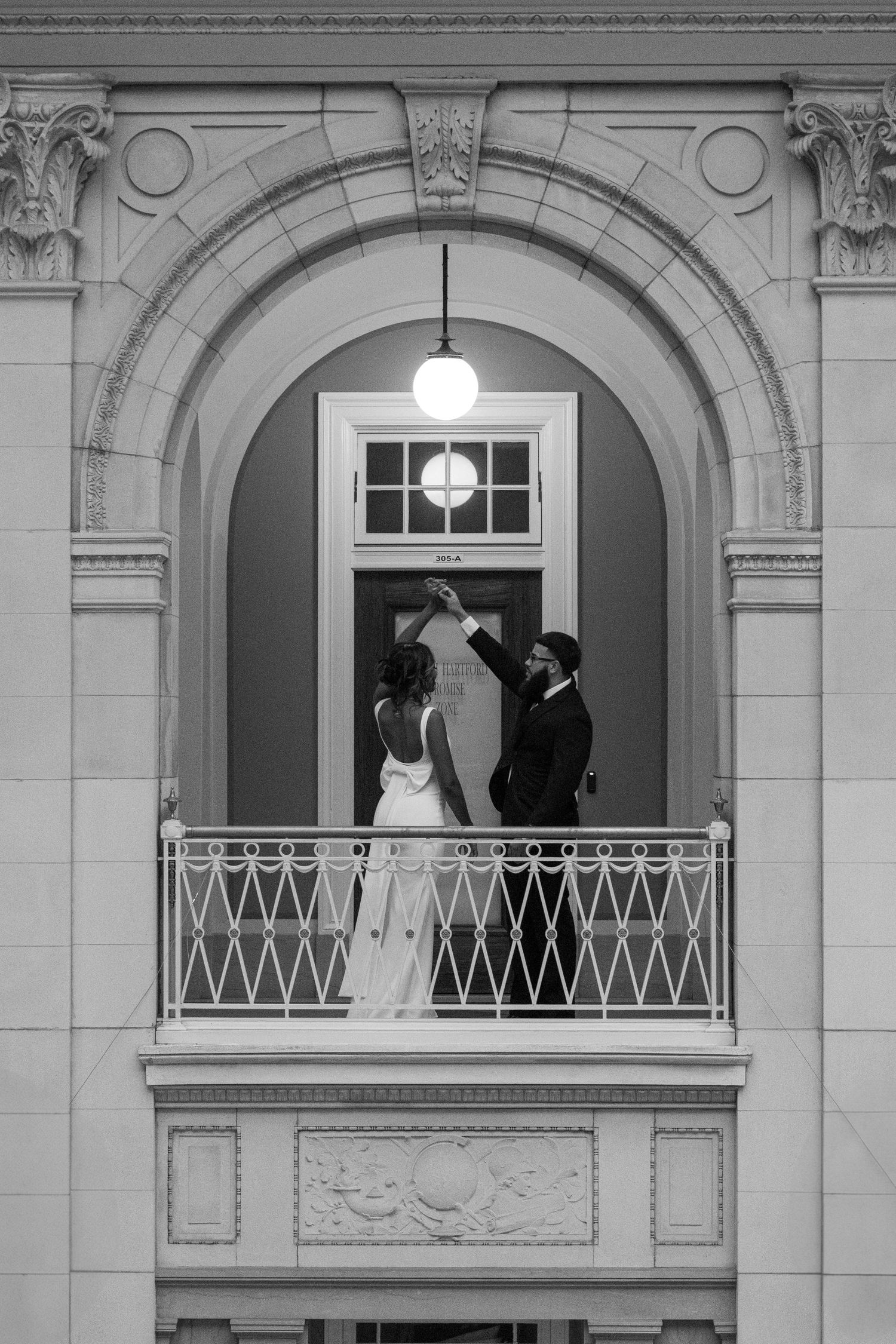 Black and white photo of bride and groom dancing on ornate balcony arch.