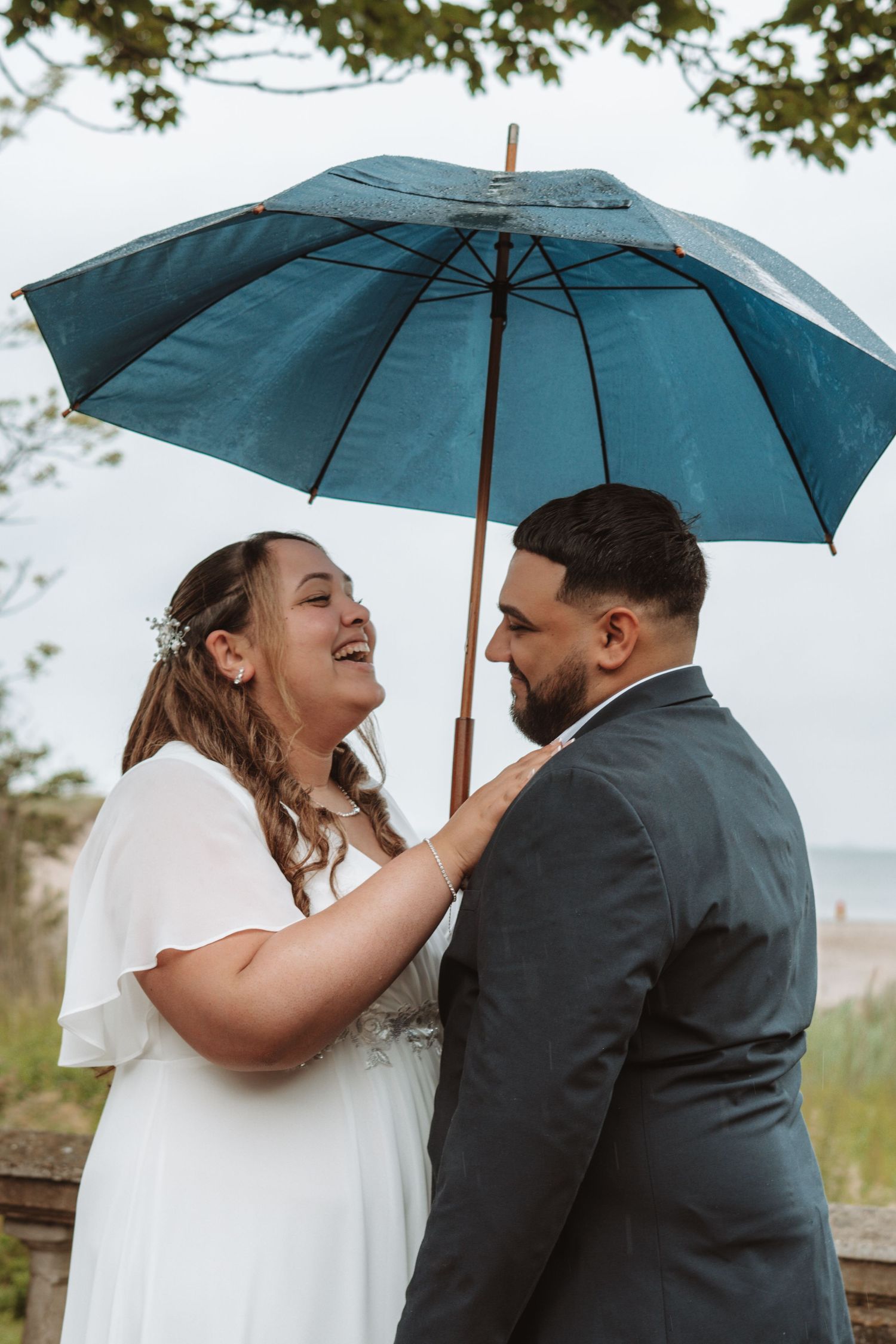 Laughing bride and groom sharing a romantic moment under a teal umbrella at their outdoor beach wedding.