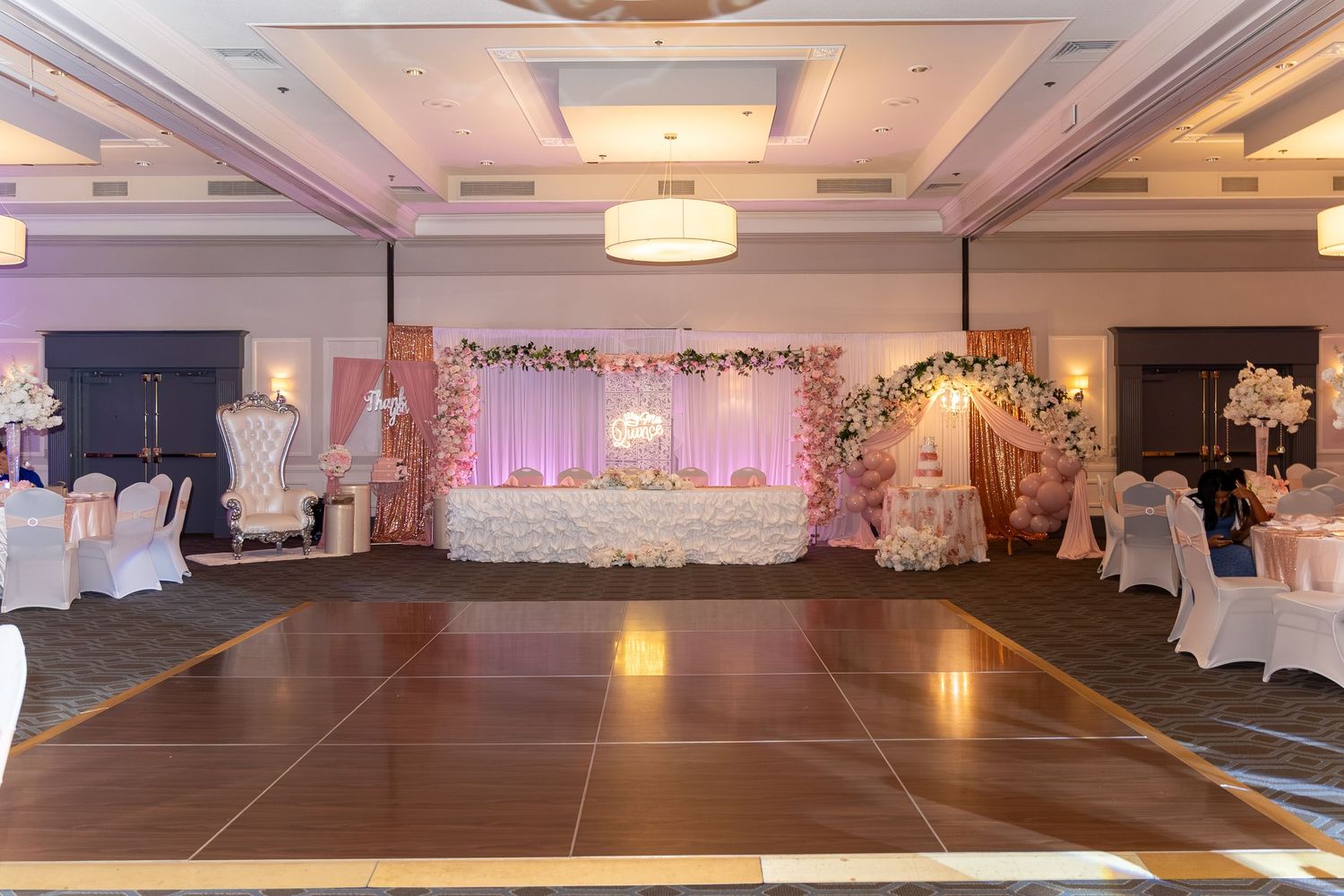 Elegant wedding reception ballroom with pink floral arch, white draped sweetheart table, dance floor, and decorated chairs. Sweet 16/Quincea