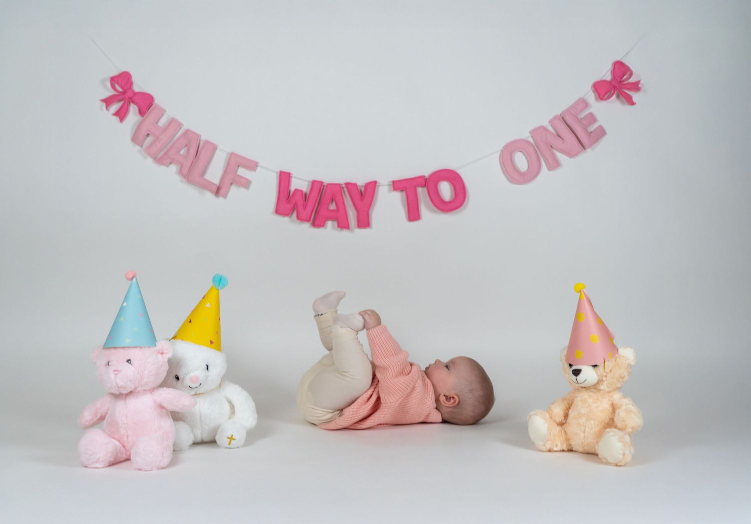 Baby lying under pink Half Way to One banner with stuffed animals and party hats.