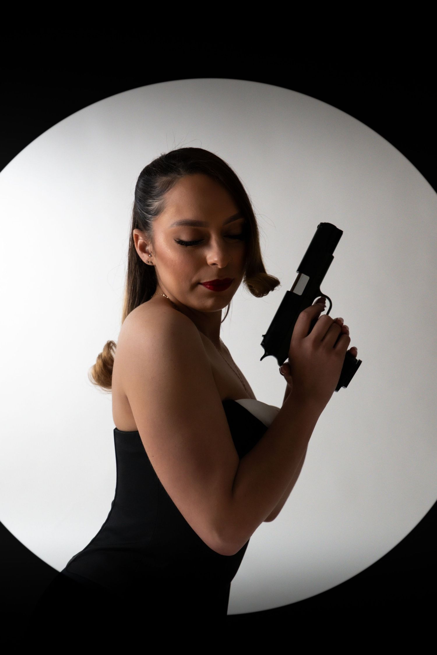 Woman in black dress holding a handgun, posed dramatically in a circular spotlight on white background.