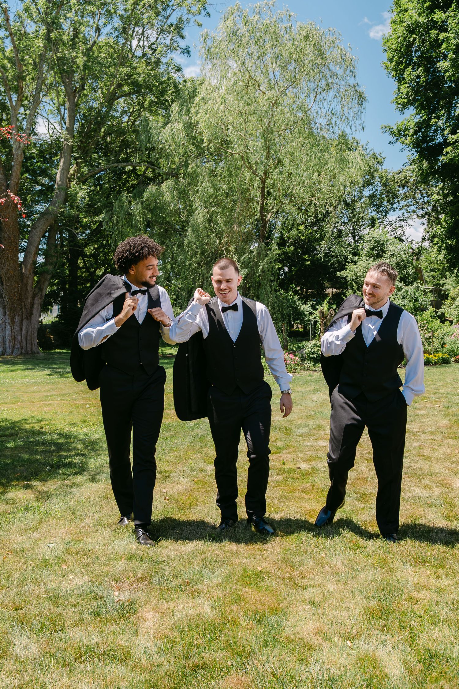 Three groomsmen in black vests and bow ties laugh while walking across a sunny green lawn at an outdoor wedding.