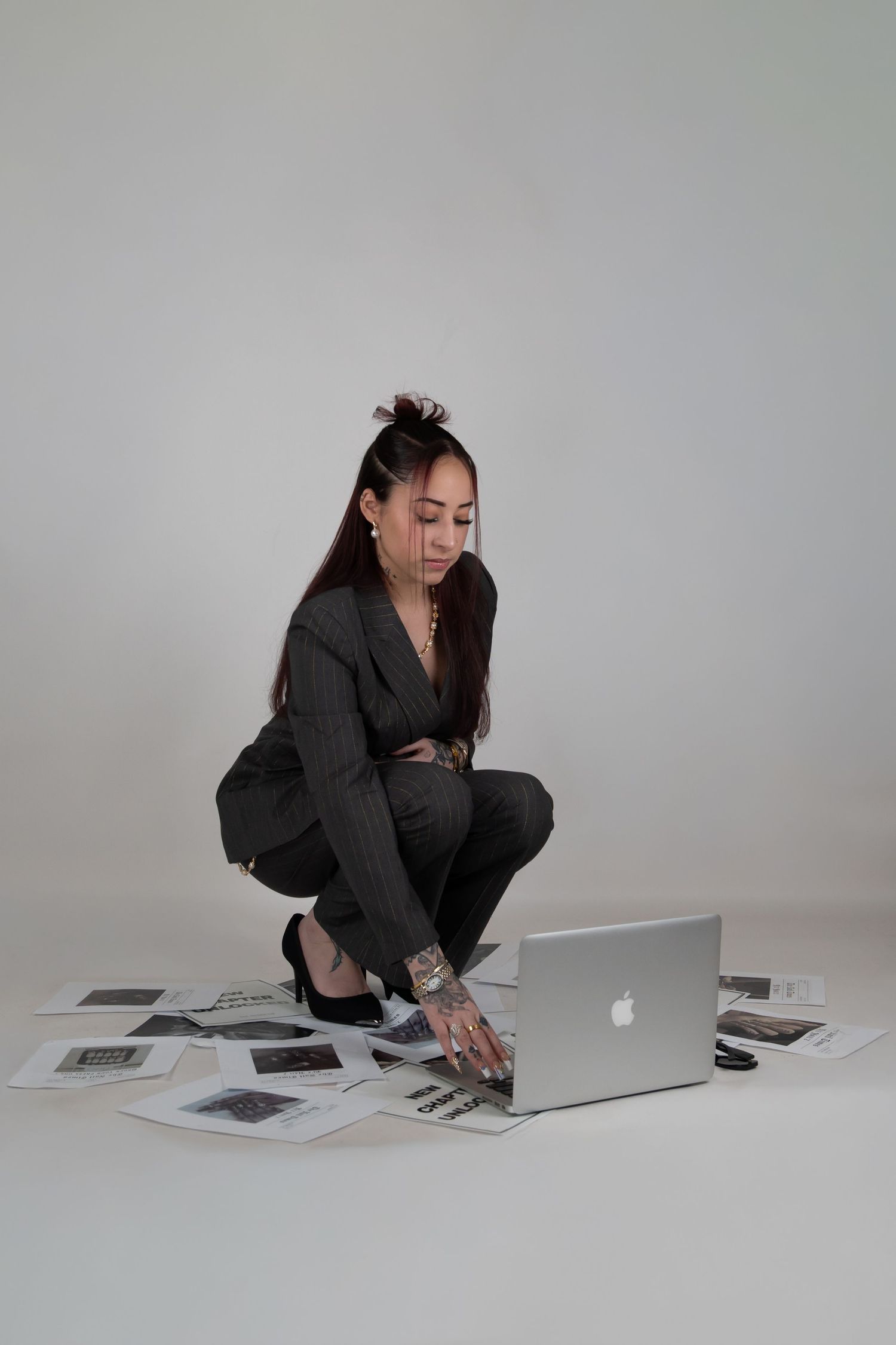 Woman in gray suit crouching on floor with Apple laptop surrounded by printed photos in studio setting. Business photoshoot in Connecticut