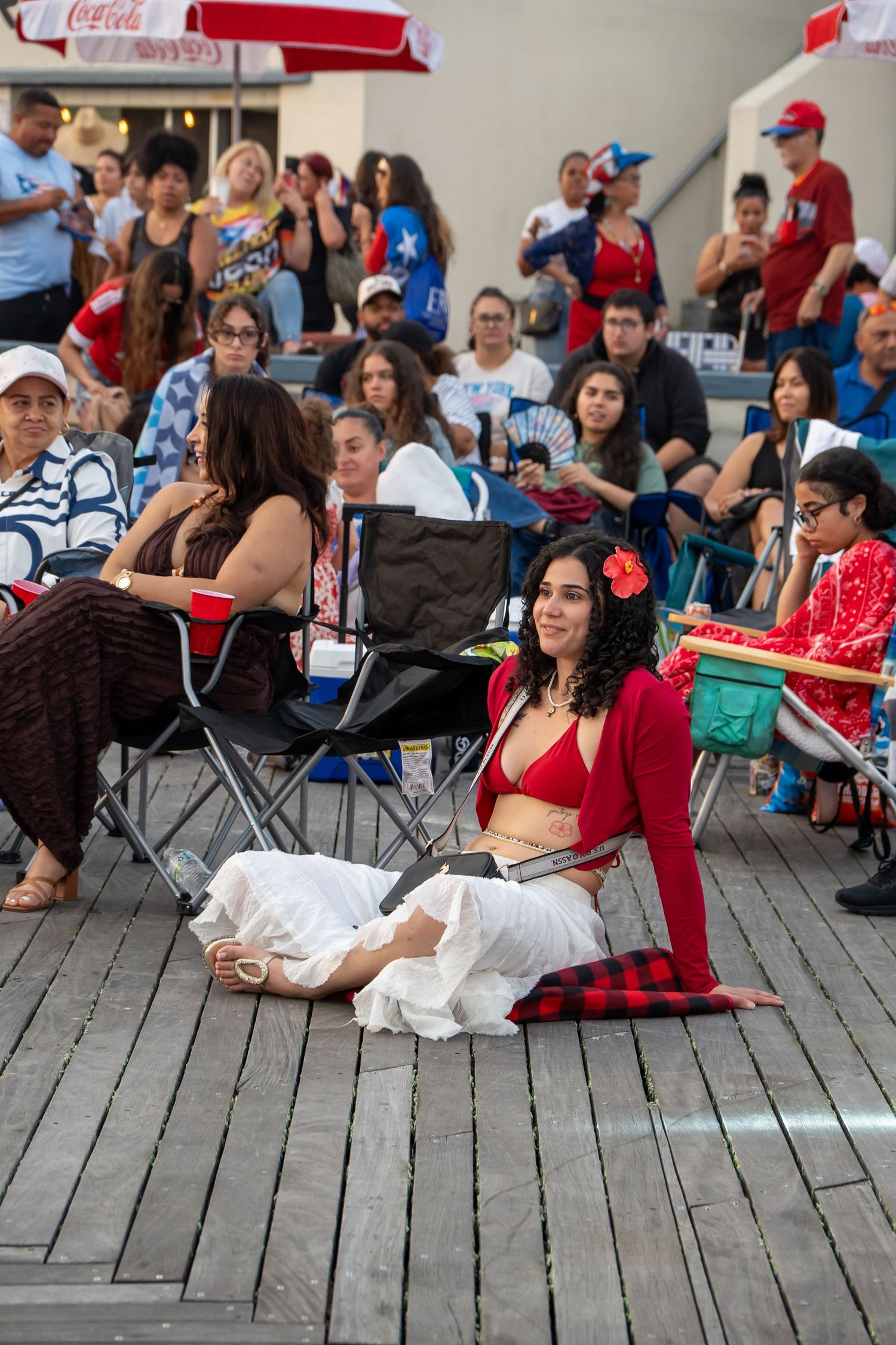 Woman in red bikini top and white skirt sits on wooden boardwalk at outdoor event with crowd behind her.