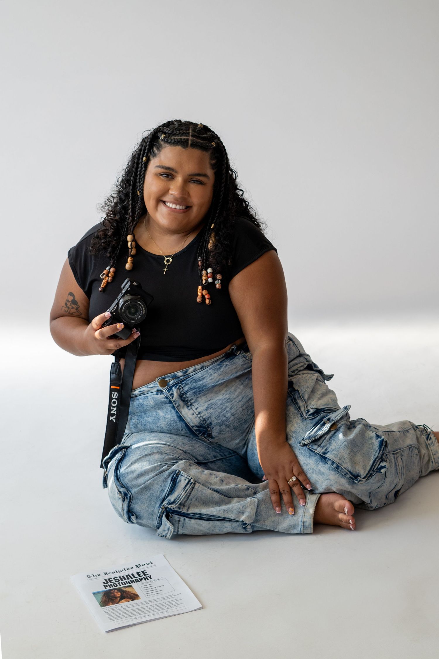 Smiling woman sitting on floor holding Sony camera, wearing black crop top and ripped jeans, with curly hair and beaded earrings.