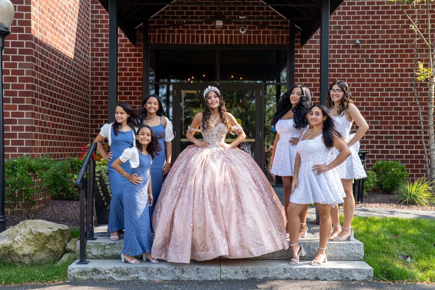 A quinceañera girl in a pink ball gown poses with her court of honor in white and blue dresses outside a brick building. Sweet 16/Quinceañer