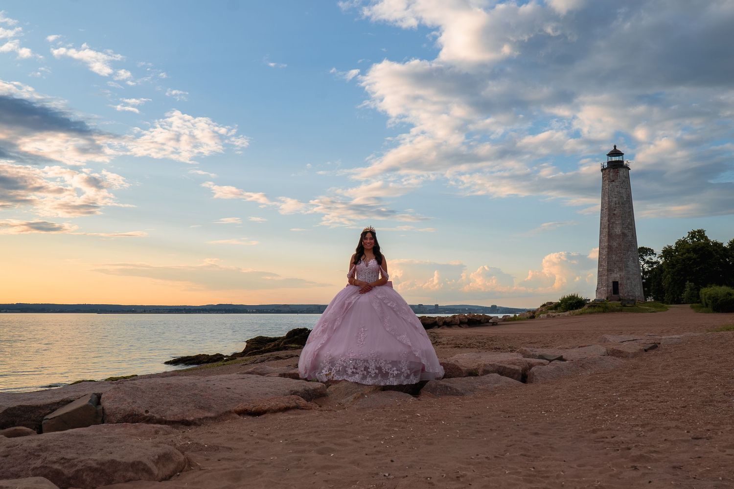Girl in pink ball gown poses near lighthouse at sunset by the water on rocky shore.
