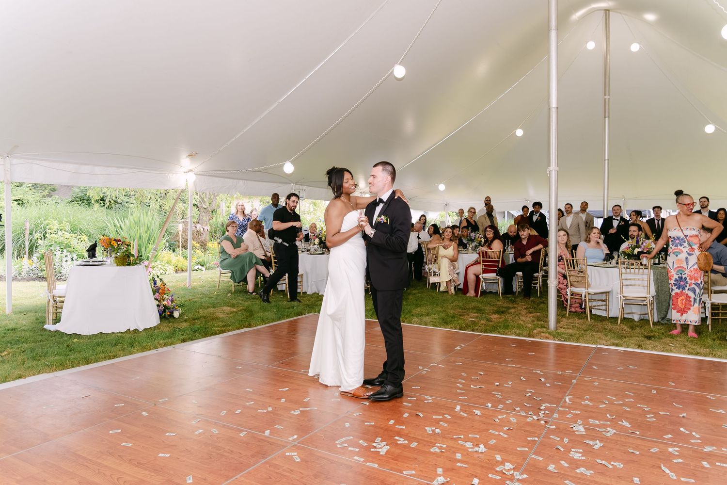 Bride and groom share first dance under white tent at outdoor wedding reception with guests seated around dance floor.