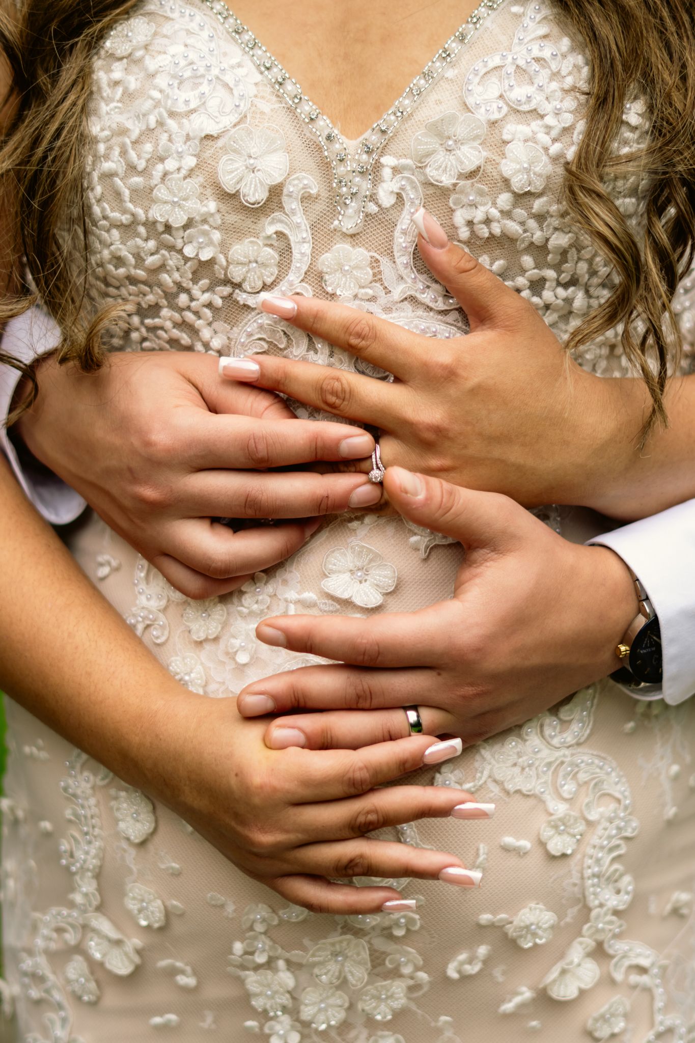 Close-up of bride and groom hands showing wedding rings on lace bridal gown.