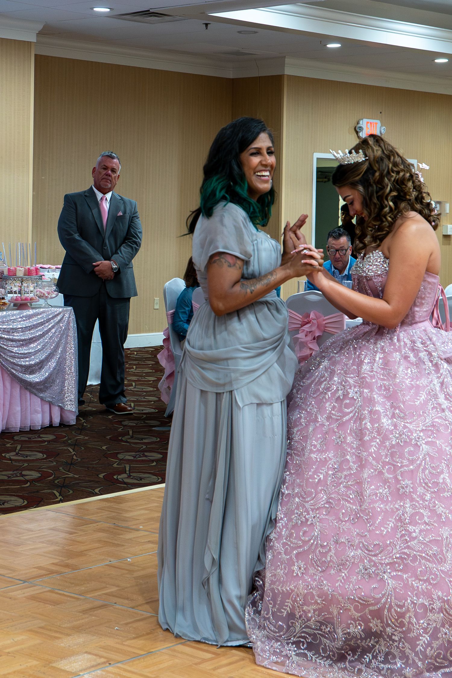 Woman in gray dress laughing with girl in pink quinceañera gown at elegant celebration while man in suit watches. Sweet 16!