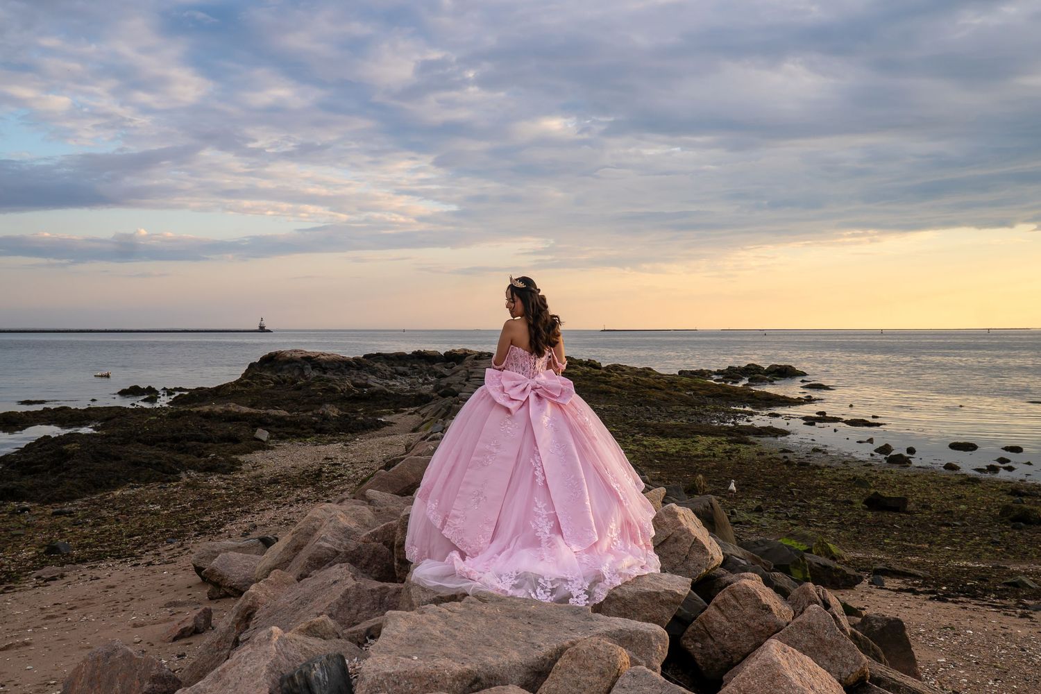Woman in pink ball gown standing on rocky shore at sunset with ocean and lighthouse in background.
