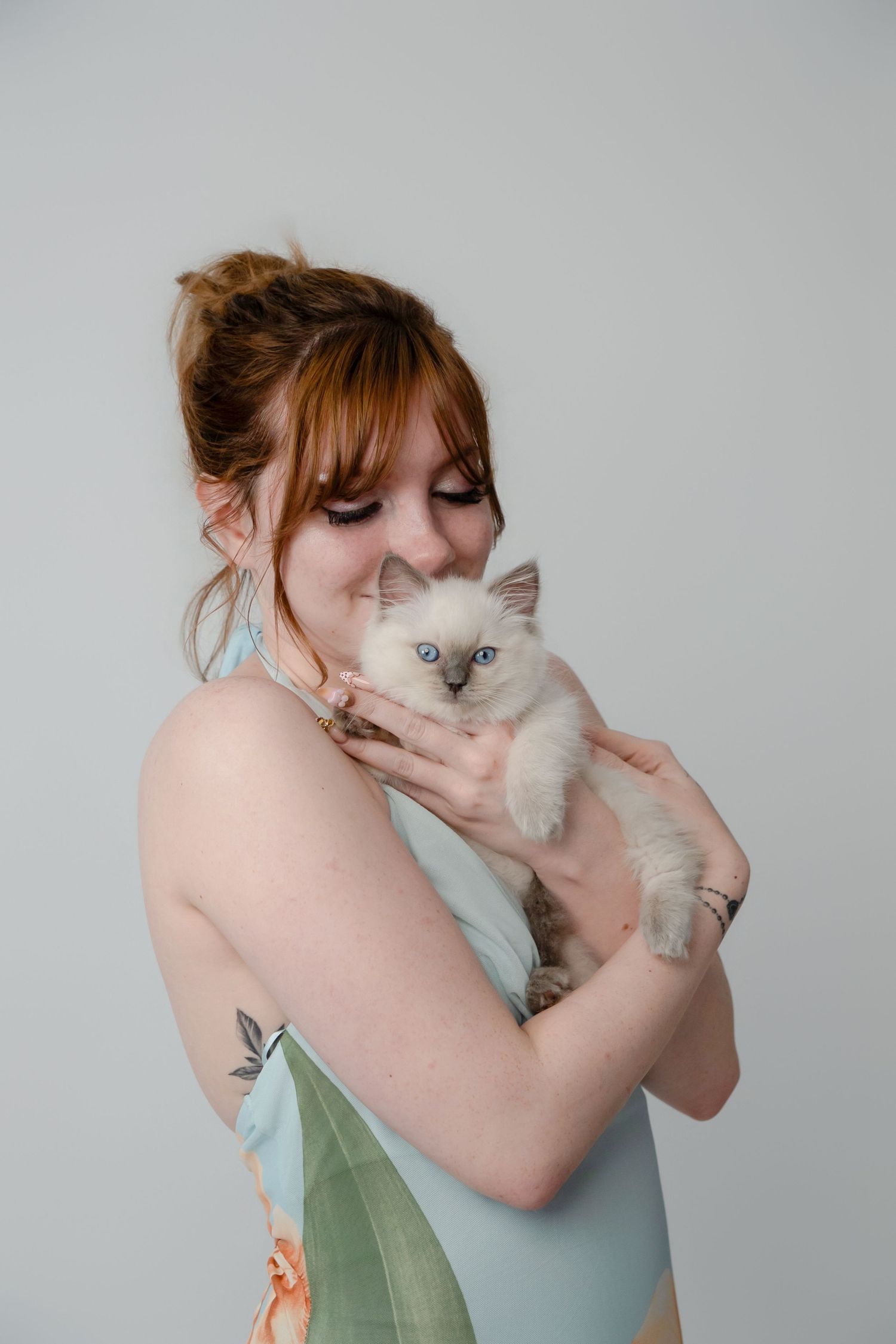 Red-haired woman in blue dress holding a white Ragdoll kitten with blue eyes against a gray background.