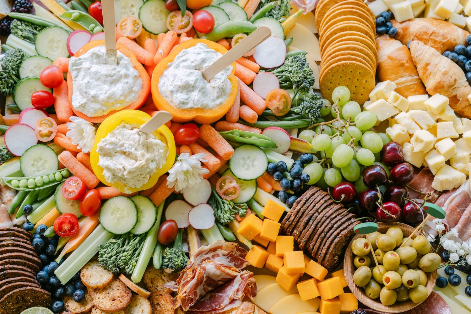 A colorful charcuterie board with fresh vegetables, fruits, cheeses, crackers, dips, and olives arranged beautifully.