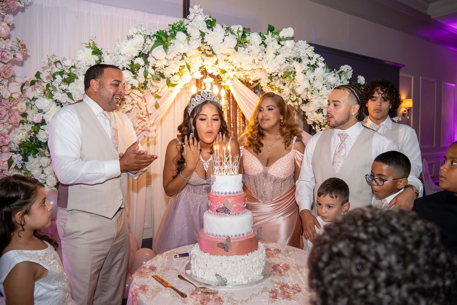 Girl blows out candles on pink birthday cake at quinceañera surrounded by family. Sweet 16/Quinceañera