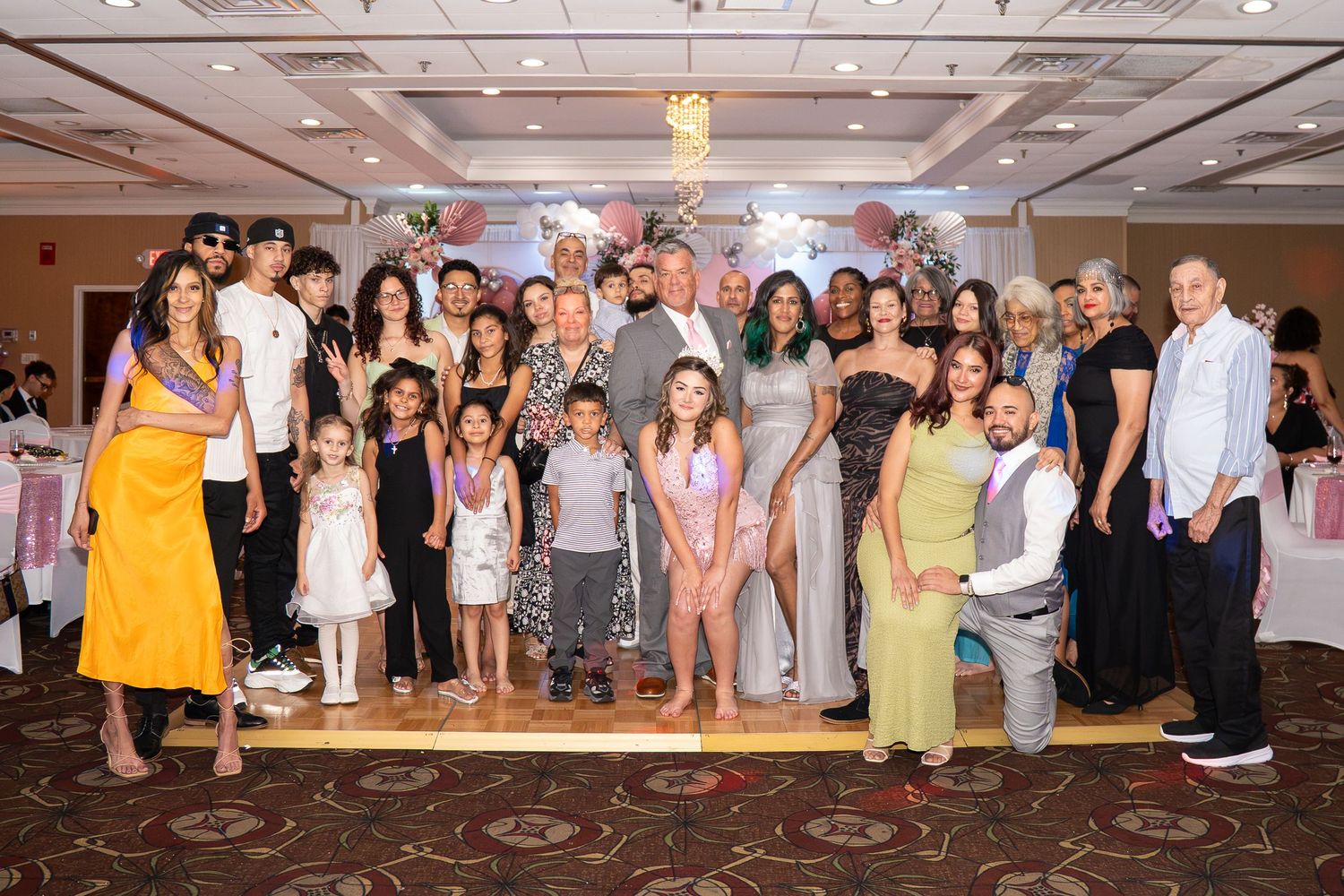 Large family group posing together at a festive indoor celebration with floral decorations in a banquet hall. Sweet 16!