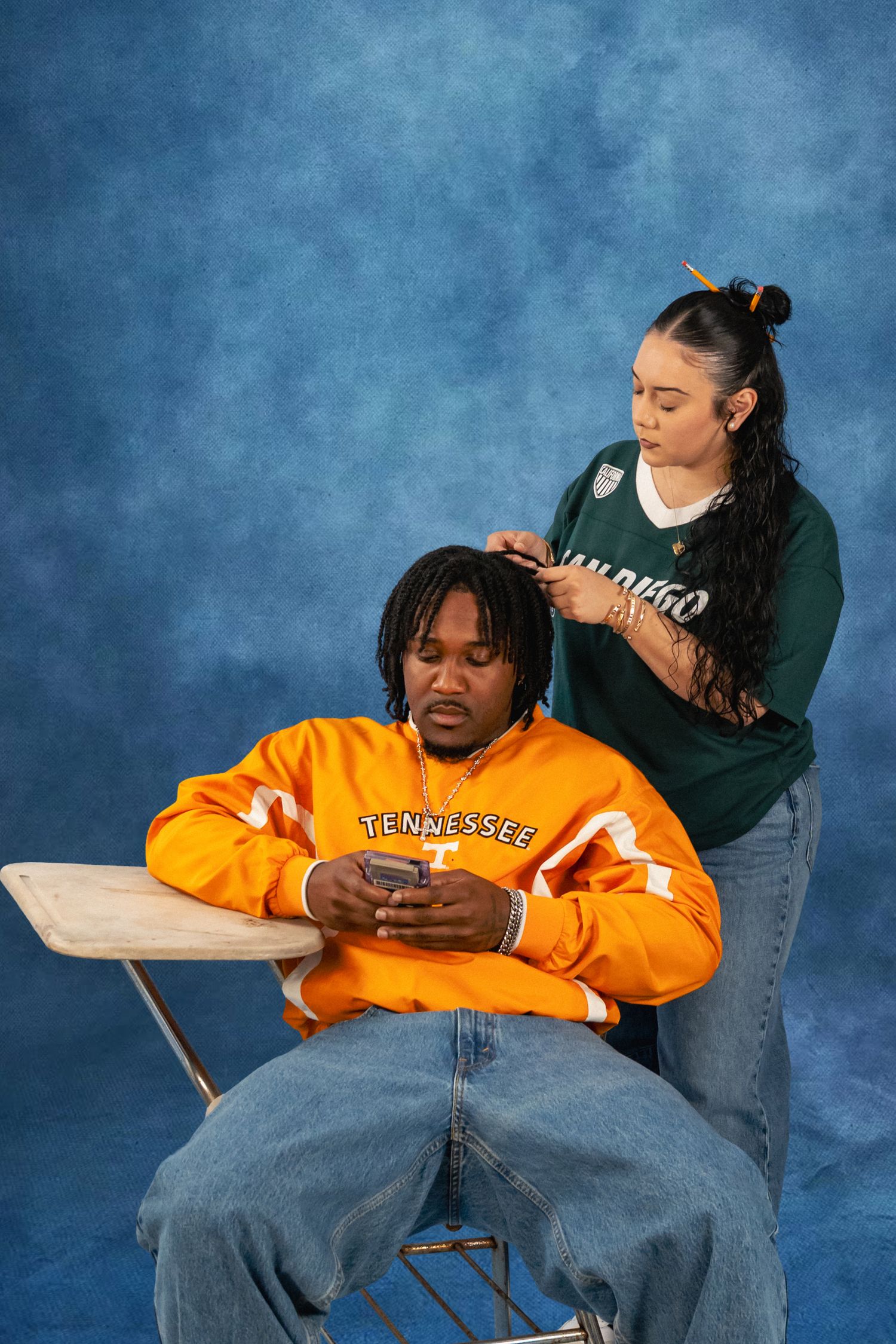 A man in an orange Tennessee sweatshirt sits in a school desk chair looking at his phone while a woman in a green jersey styles his hair.