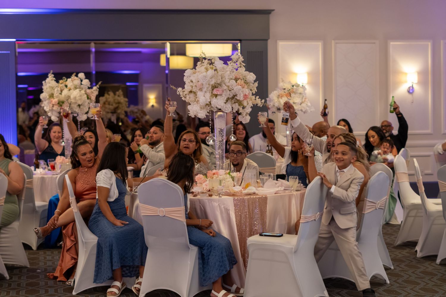Joyful wedding guests raising hands at decorated reception tables with tall floral centerpieces. Sweet 16!