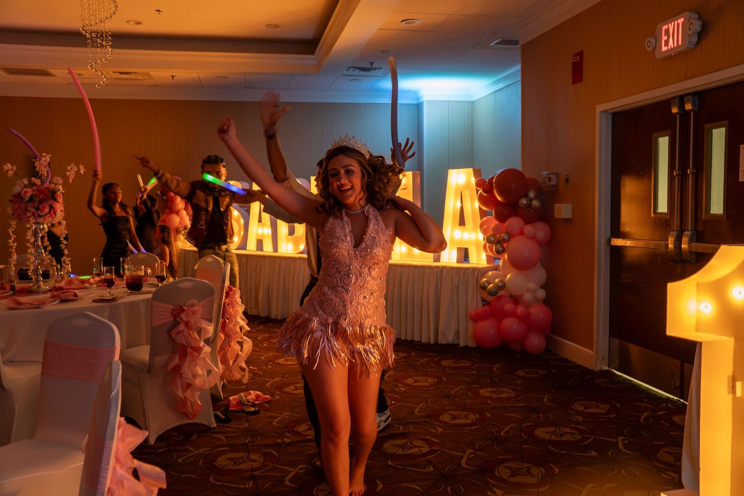 Woman in feathered costume dancing at a festive party venue with balloon decorations and marquee letters. Sweet 16!