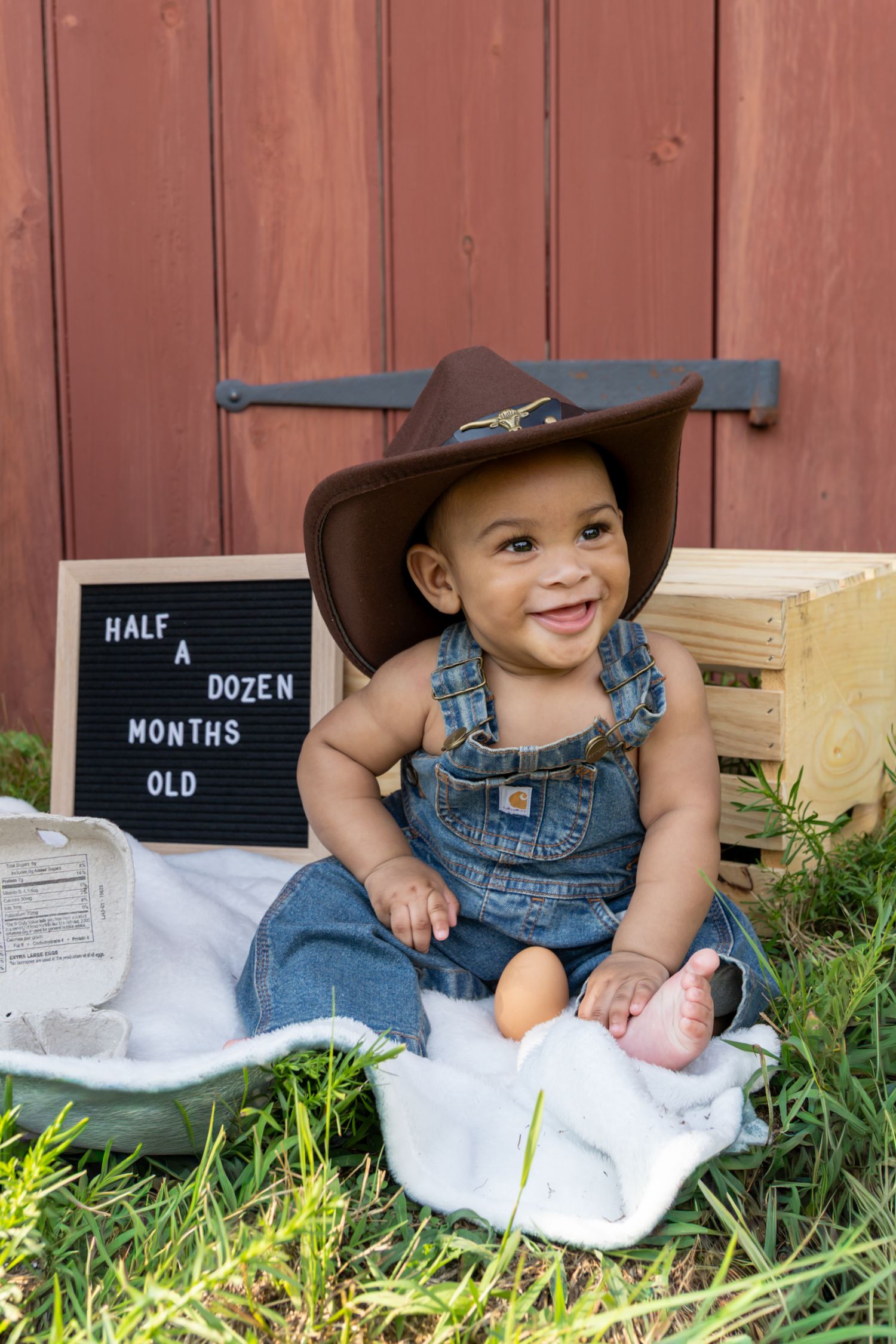 Smiling baby in cowboy hat and denim overalls sits outdoors beside a sign reading Half A Dozen Months Old. 6 month photoshoot