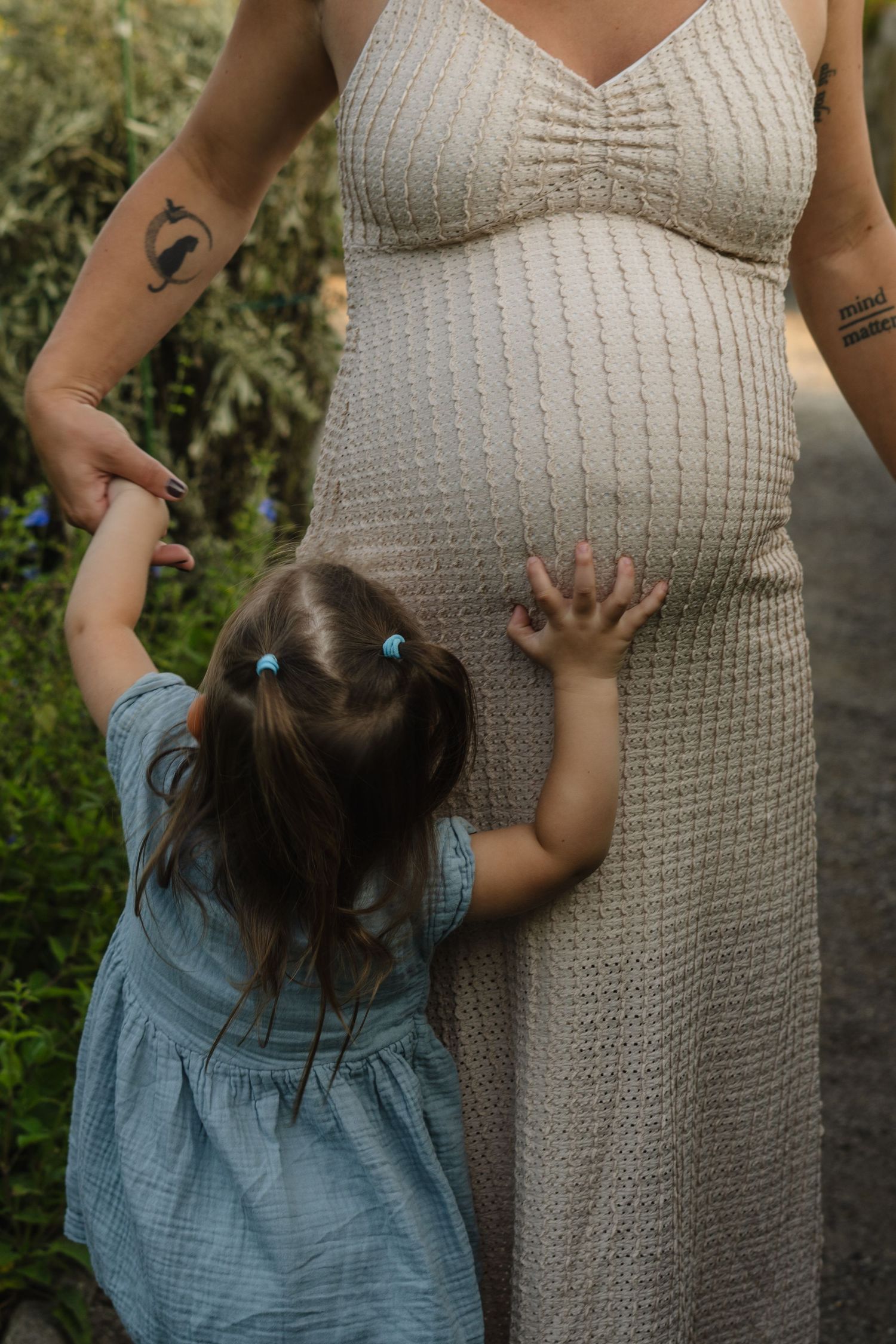 Young girl touching pregnant mothers belly outdoors in garden setting.