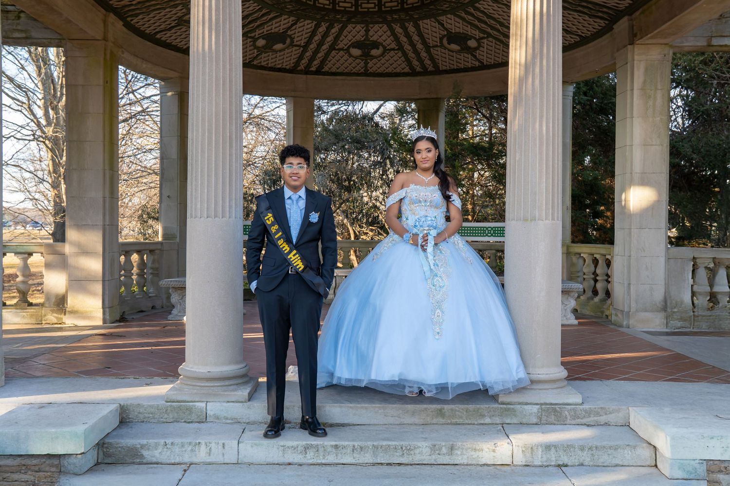 Teen couple poses in formal attire at stone gazebo for quinceañera celebration outdoors.
