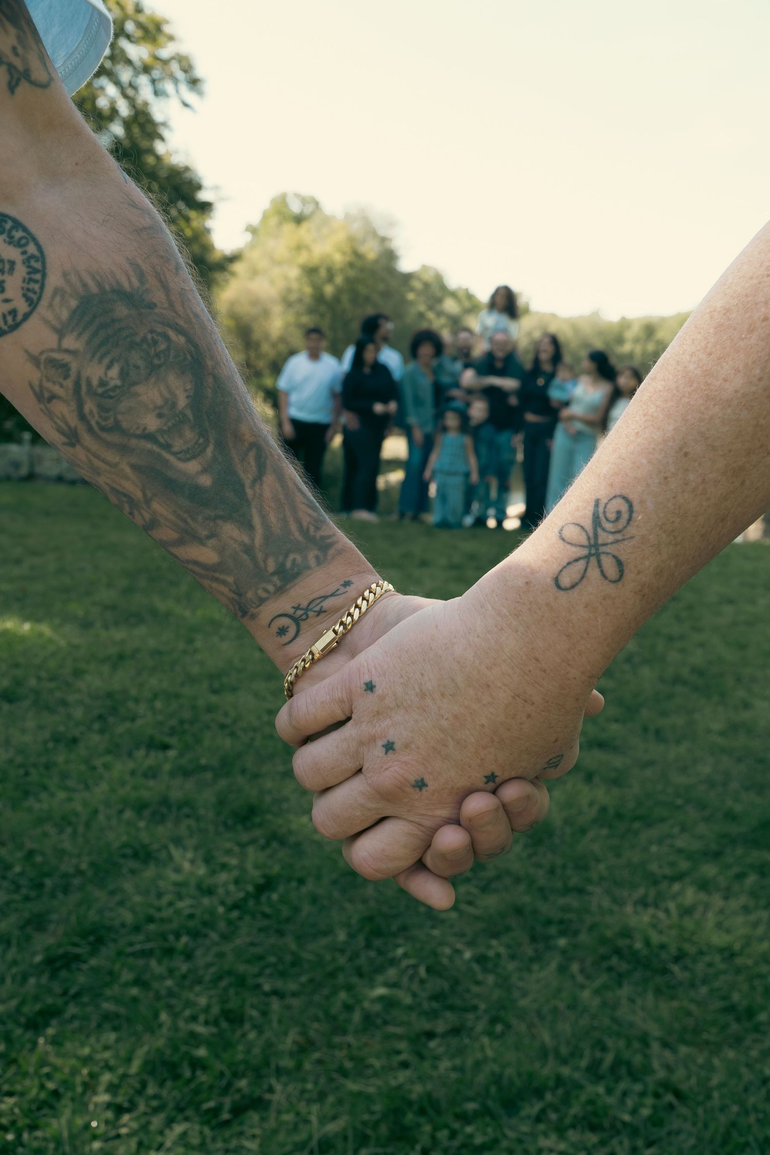 Two tattooed hands clasped together in the foreground on a grassy lawn, with a large family group blurred in the background.